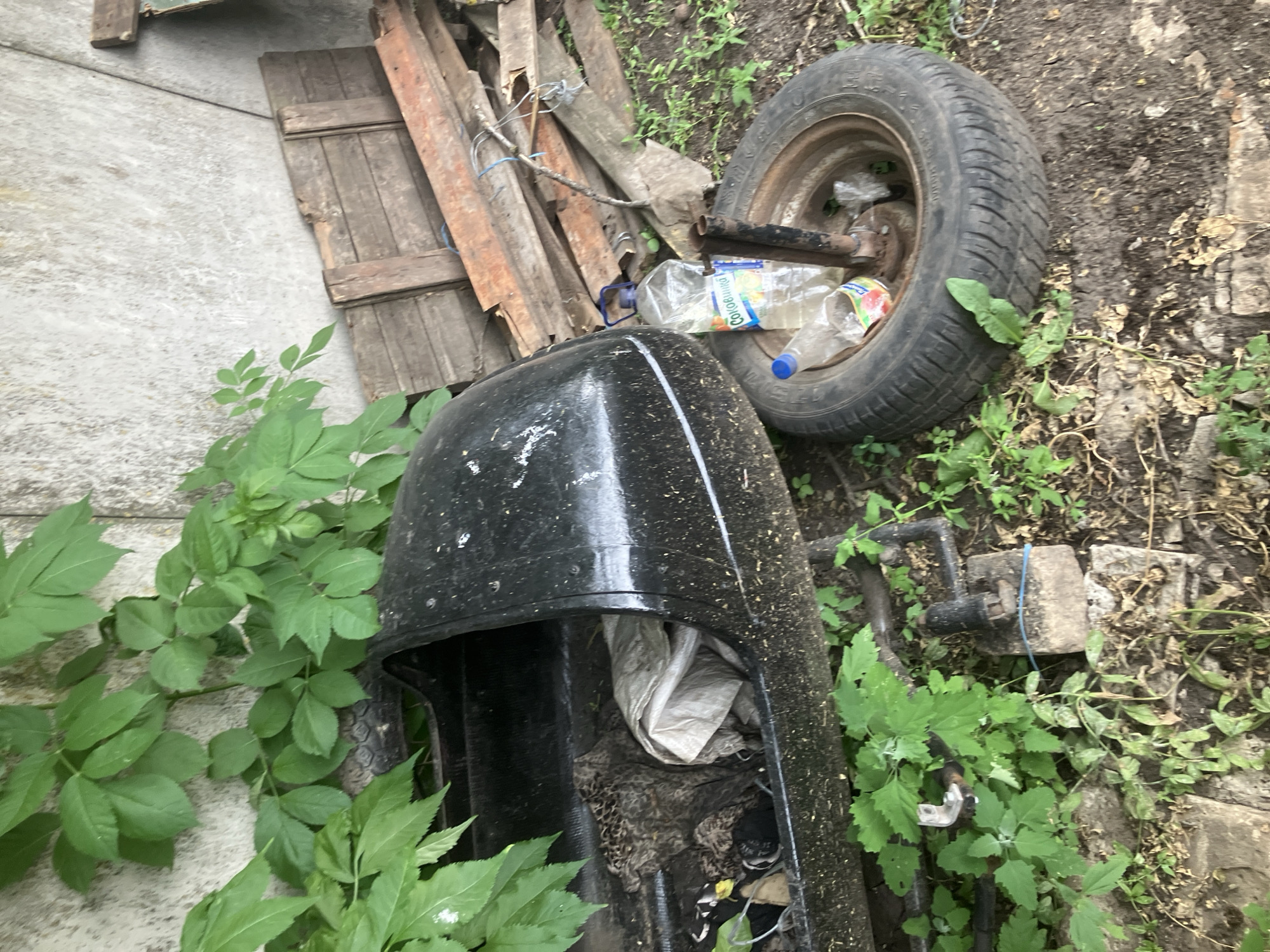 A dusty, black plastic object, possibly a car roof box, lies on its side amidst overgrown vegetation next to a concrete wall in Vovkivka, Ukraine. A spare tire is positioned nearby, its rim showing signs of rust. Two plastic bottles, one partially filled with a clear liquid and bearing a label with Cyrillic text, are placed on the tire. Scattered wooden planks are stacked against the wall in the background. The ground is covered with dirt, dried leaves, and green plants, indicating an outdoor, possibly neglected, setting. No people are present in the scene.