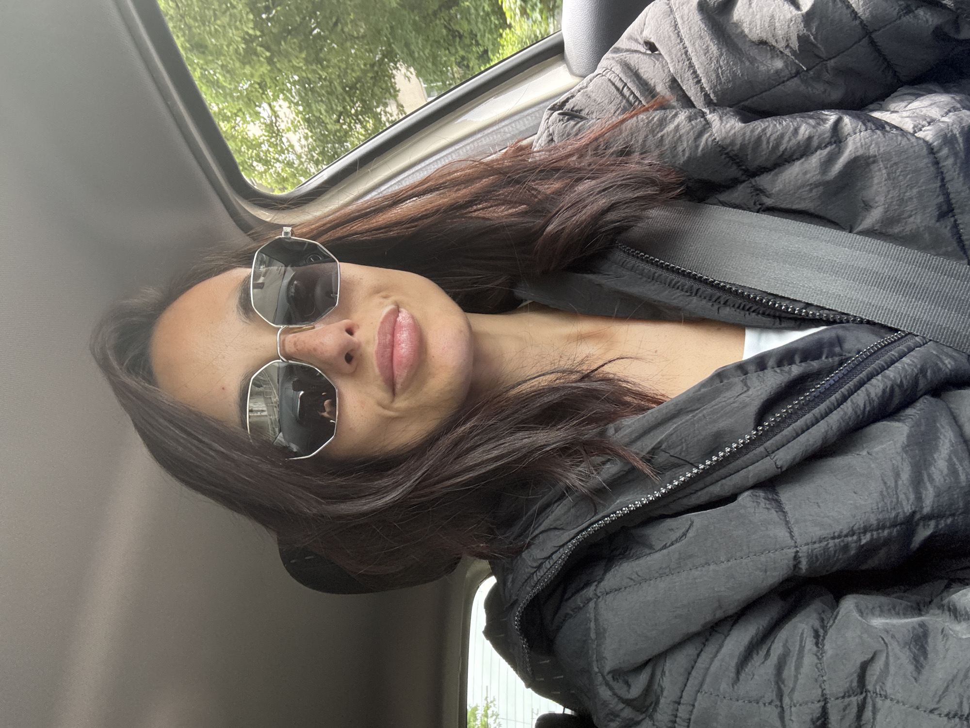 The image shows a woman wearing sunglasses and a black quilted jacket with a seatbelt. She is inside a car. The view outside the car window suggests an urban environment with trees. It is not possible to identify the location as Pleven, Bulgaria, or any other specific location, based on the information in the image.