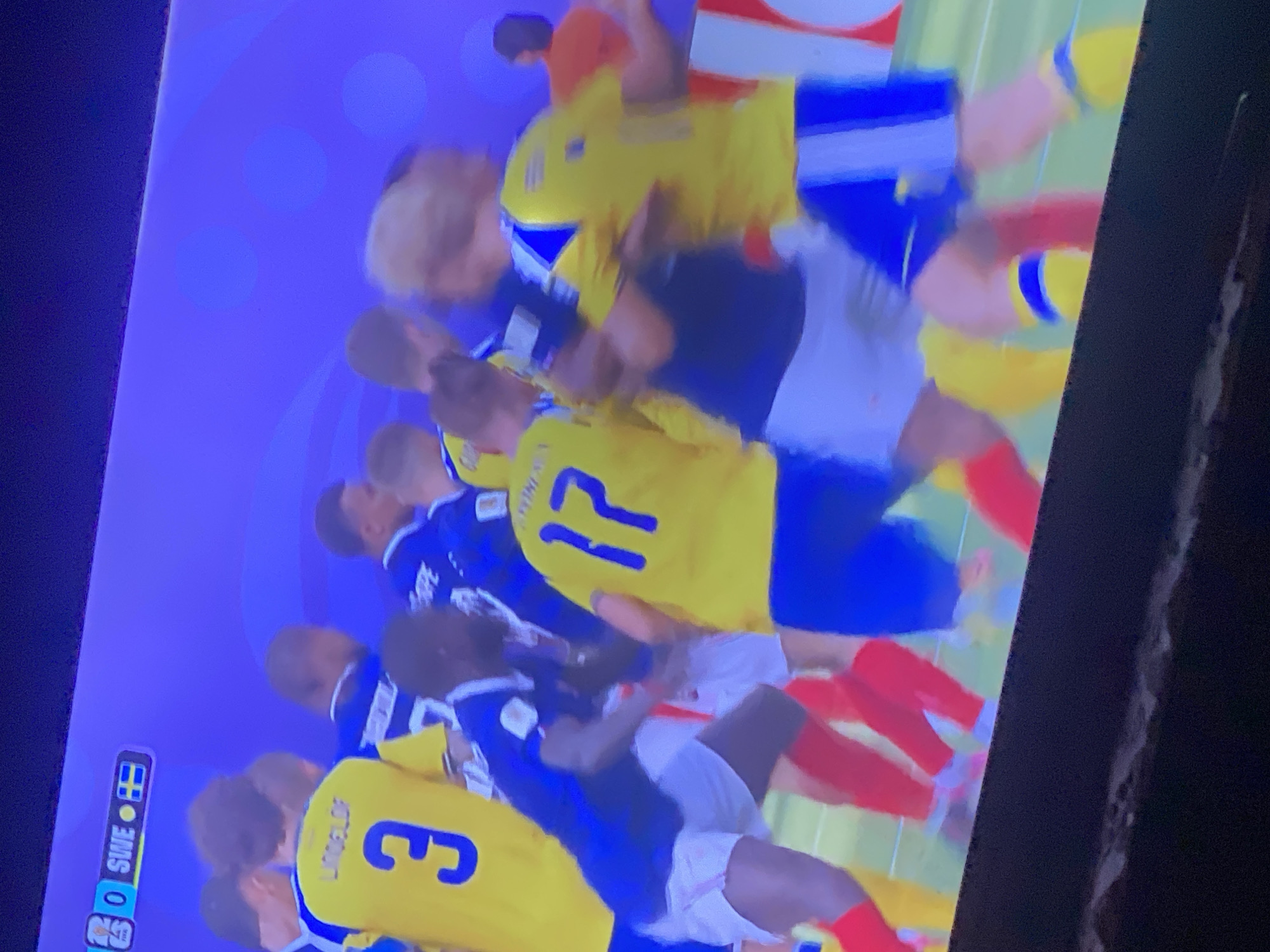 The image displays a soccer match, as indicated by the score and team names visible in the upper left corner: "0 SWE 0". Players in yellow and blue jerseys are engaged in gameplay. One player in a yellow jersey with the number "3" and the name "LINDELOF" on the back is partially visible. Another player in a yellow jersey with the number "17" is also present. Players in blue jerseys appear to be on the opposing team. The background is a solid purple, suggesting a digital or projected environment, not a real-world stadium in Zastavna, Ukraine. The scene captures a moment of physical interaction between players, typical of a soccer game.
