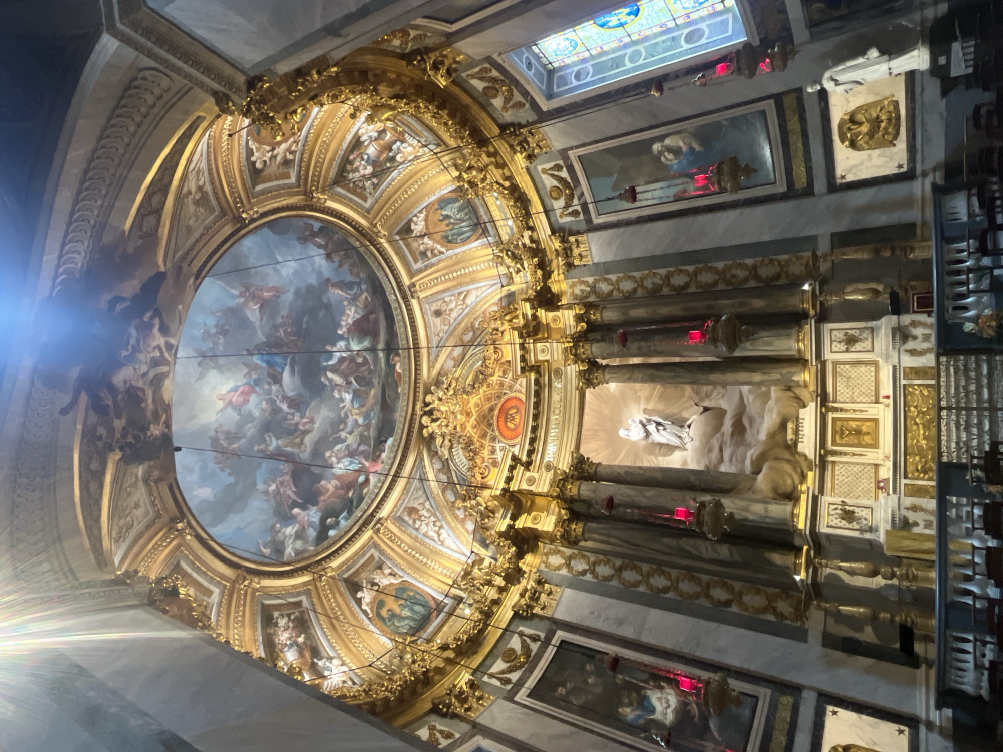 The image depicts the interior of the Église Saint-Roch in Paris, France. The view is angled upwards, focusing on the ornate altar area and the ceiling. The altar is a grand structure with classical columns and a large, detailed sculptural group in white marble, depicting a mother and child figure ascending on clouds, surrounded by ornate gold detailing. Above the altar, a large circular fresco adorns the ceiling, depicting a celestial scene with numerous figures, possibly religious in nature, rendered in blues and browns.
Flanking the altar are large paintings, one on each side, set within gold frames and appearing to depict religious narratives with figures in period attire. Hanging from the ceiling are several decorative pendant lights, some emitting a red glow. Stained glass windows, featuring intricate patterns and vibrant colors, are visible on the right side of the image, allowing light to filter into the space. The walls are clad in a marble-like material, with decorative gold trim and various framed artworks and sculptures integrated into the architecture. In the foreground, a railing separates the viewer from a section of seating, likely pews, indicating a place of worship. The overall impression is one of opulence and religious grandeur, characterized by rich ornamentation, intricate artwork, and dramatic lighting.