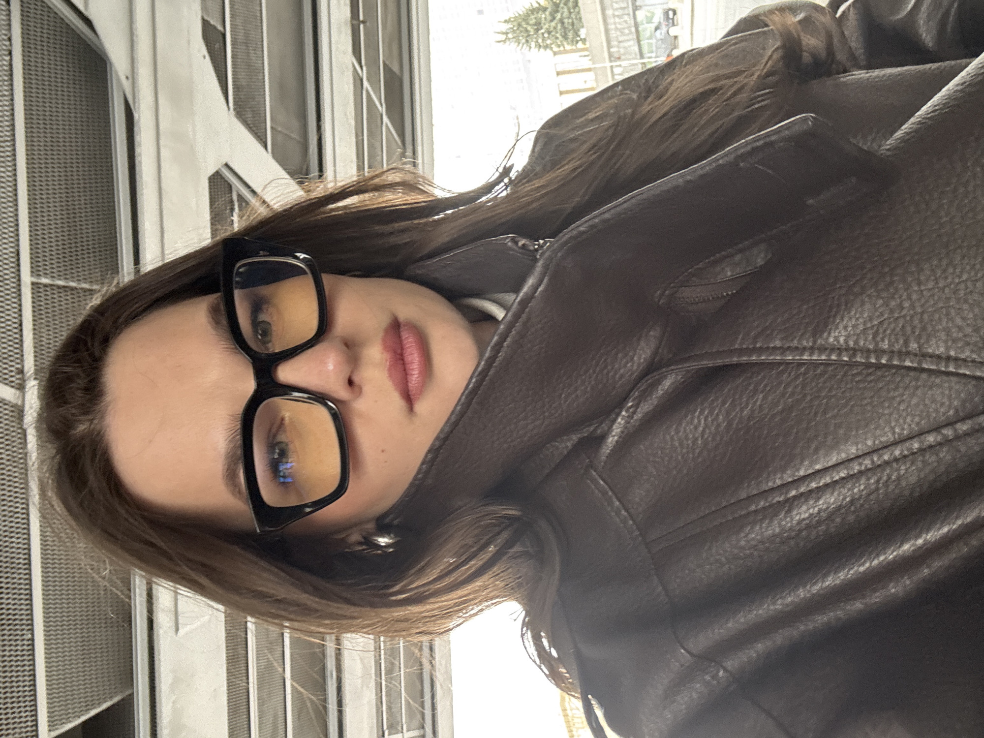 A woman with brown hair, wearing glasses and a brown leather jacket, is pictured in a close-up selfie. She is standing in front of a building structure made of white steel beams and a mesh-like wall. Outside, blurred in the background, a tree and other buildings suggest an urban environment. There is no concrete information in the photo to indicate this picture was taken in Moscow, Russia.