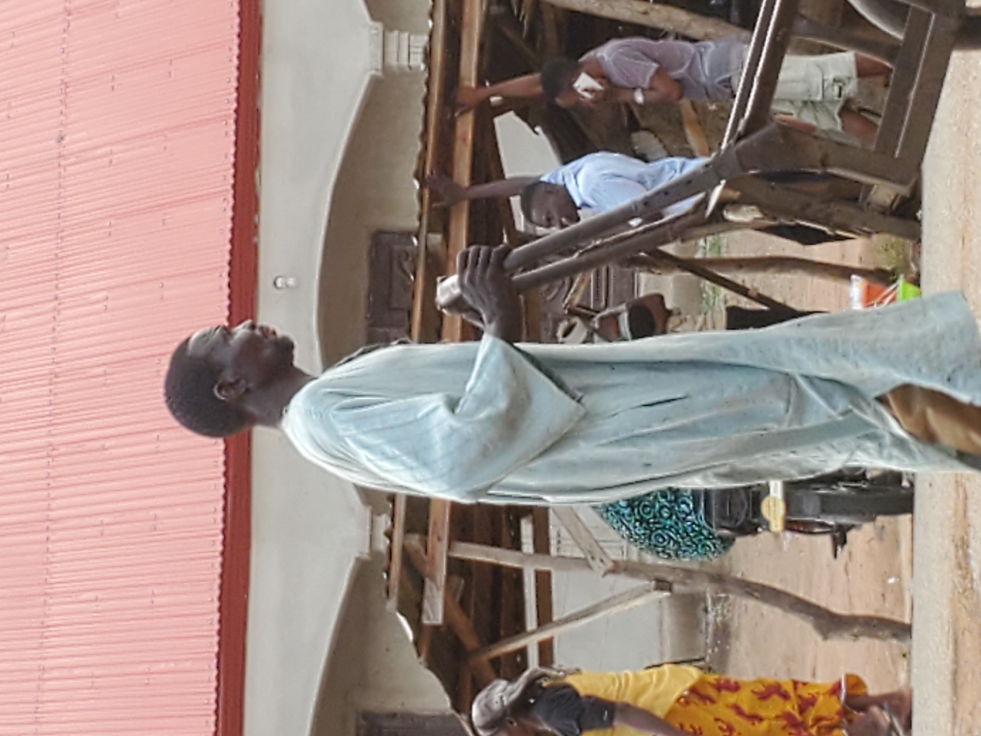 The image depicts an outdoor or semi-outdoor scene, likely a construction site or an area under renovation, featuring several individuals and wooden structural elements. The perspective suggests the image is rotated 90 degrees clockwise; when oriented correctly, a prominent red corrugated metal roof is visible along the top.
In the foreground, on the right side, a man with dark skin and short dark hair is standing, looking upwards and slightly to his left. He is wearing a light blue or greenish-blue long-sleeved tunic-like garment. His right hand is holding onto a dark, possibly metal, pole or part of a piece of equipment.
In the midground, several other people are visible amidst a complex framework of wooden beams and supports. One man, wearing a light blue short-sleeved shirt, extends his left arm towards a wooden beam. Above him, another man in a light purple short-sleeved shirt and light-colored shorts is reaching upwards, holding a white object that resembles a smartphone.
Further in the background, towards the bottom left, a person wearing a yellow and red patterned shirt is partially visible, and another individual is seen wearing a blue and green patterned garment or carrying a bag with such a pattern.
The setting includes unfinished or partially plastered walls. The overall activity appears to be related to building, repair, or maintenance, with individuals engaged in various tasks or observing the work. The bright, even lighting indicates that the photograph was taken during the daytime. No discernable text is visible in the image. The specific location remains unknown.