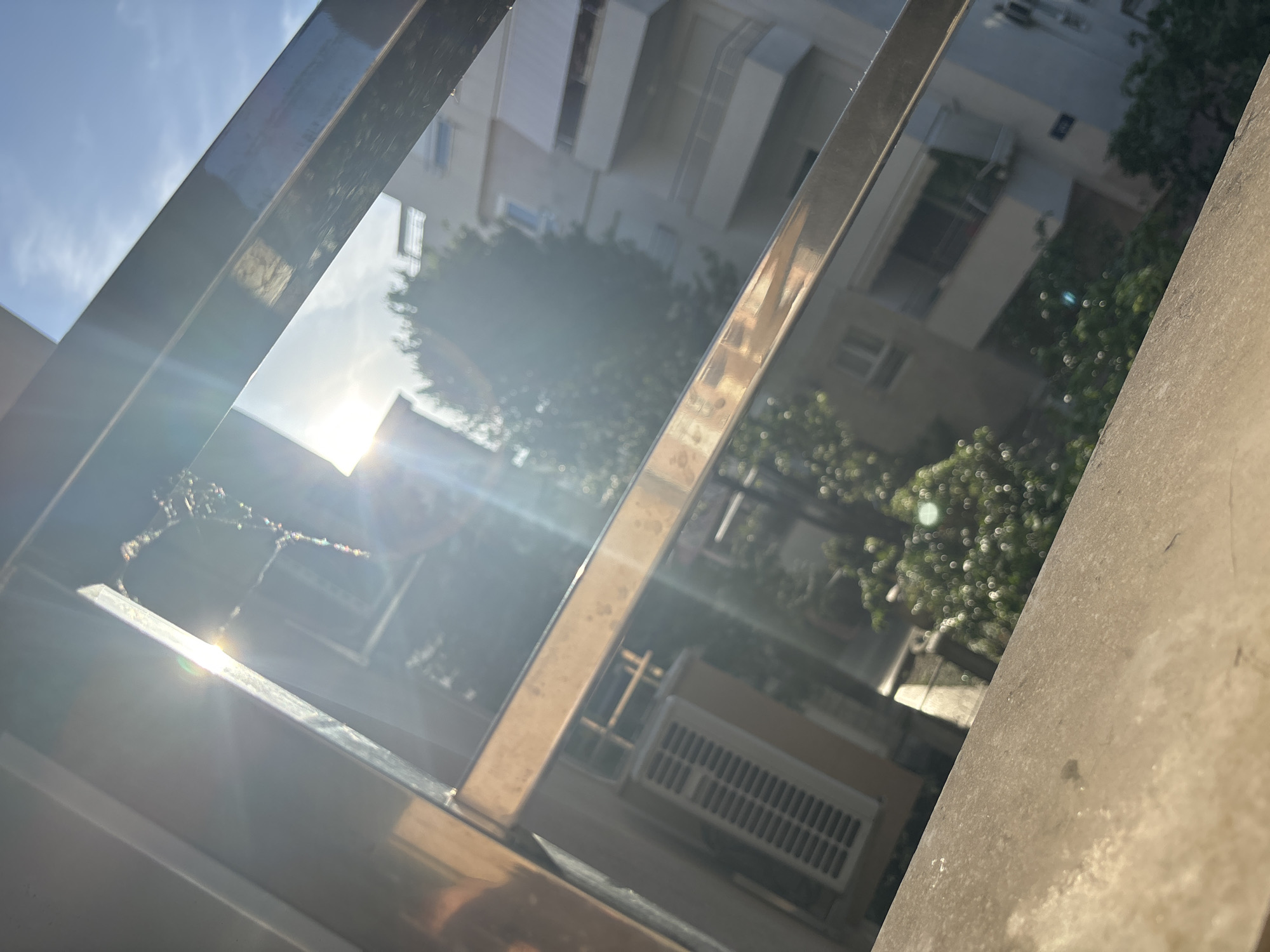 The image shows a view through a metal railing towards a building and a tree. The building appears to be residential, with multiple floors and windows. An air conditioning unit is visible on one of the floors. The tree is leafy and full. Sunlight is shining through the scene, creating lens flare. The location is possibly in Budva, Montenegro, although specific landmarks are not clear enough for confirmation. No people are visible.