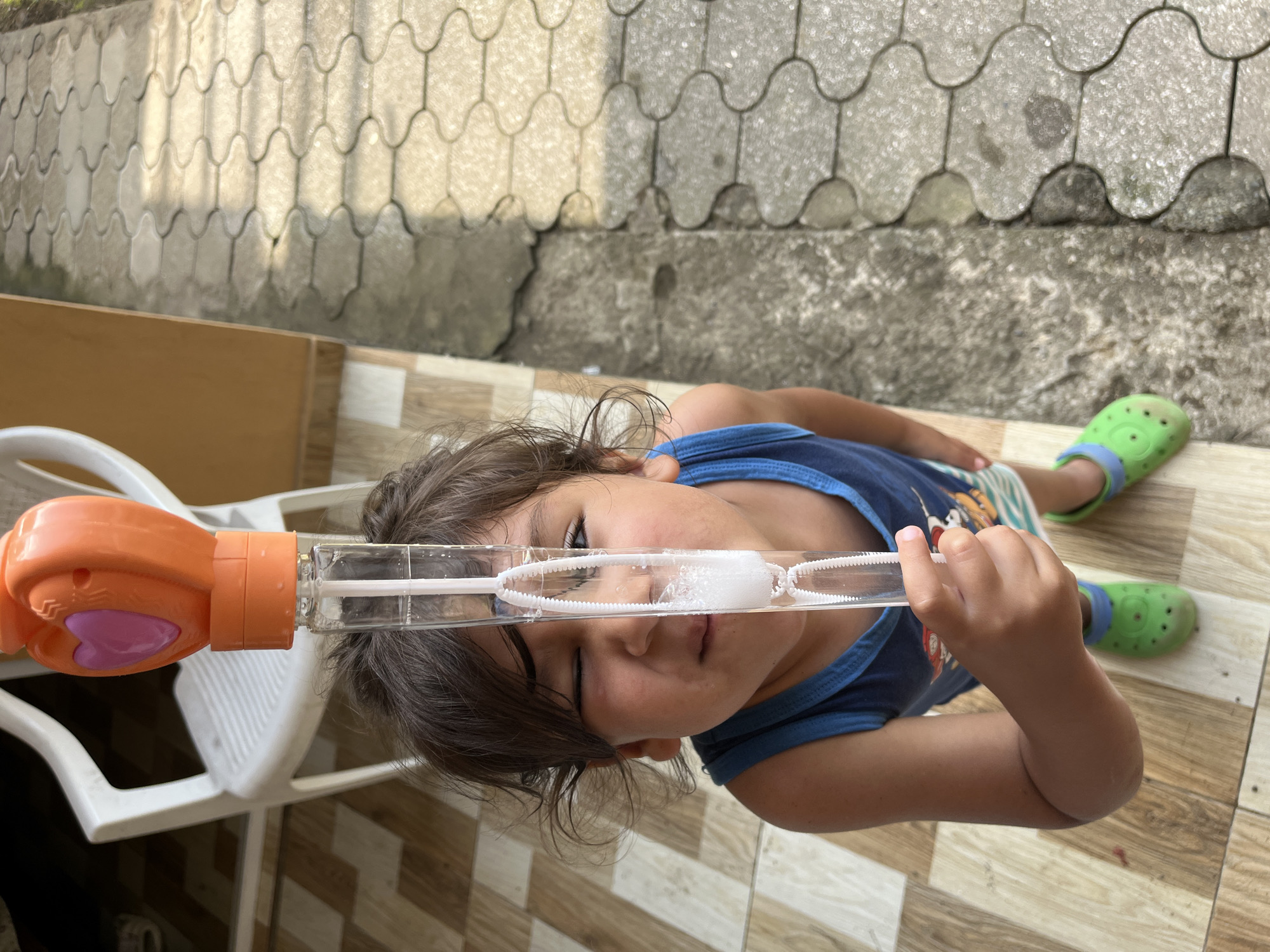 A young child stands on a tiled patio in Kobuleti, Georgia, holding a bubble-blowing toy. The child, wearing a blue tank top and green Crocs, looks up towards the toy, which is topped with an orange, heart-shaped piece. The bubble wand, filled with liquid, has several bubbles formed within it. To the left of the child, a white plastic chair is visible, and to the right, the patio transitions to a textured concrete walkway. The scene appears to be outdoors, possibly in a residential area.