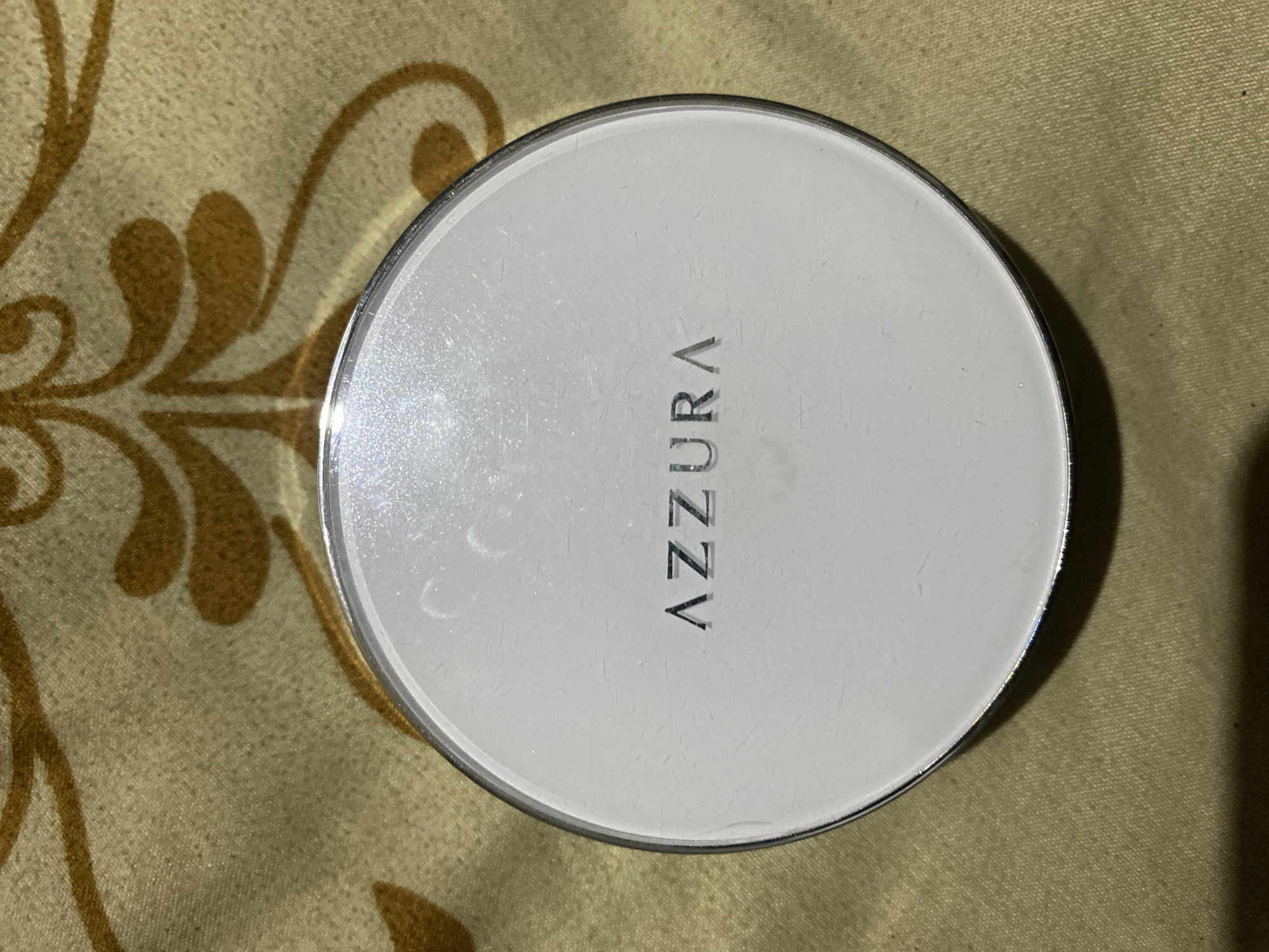 A white, circular compact powder with the brand name "AZZURA" printed in silver on its lid rests on a light brown, textured fabric with a gold ornate pattern. The compact has a silver rim. The image is a close-up, top-down shot. There are no people present, and the setting is indoors. This image is not in Ta', Indonesia.