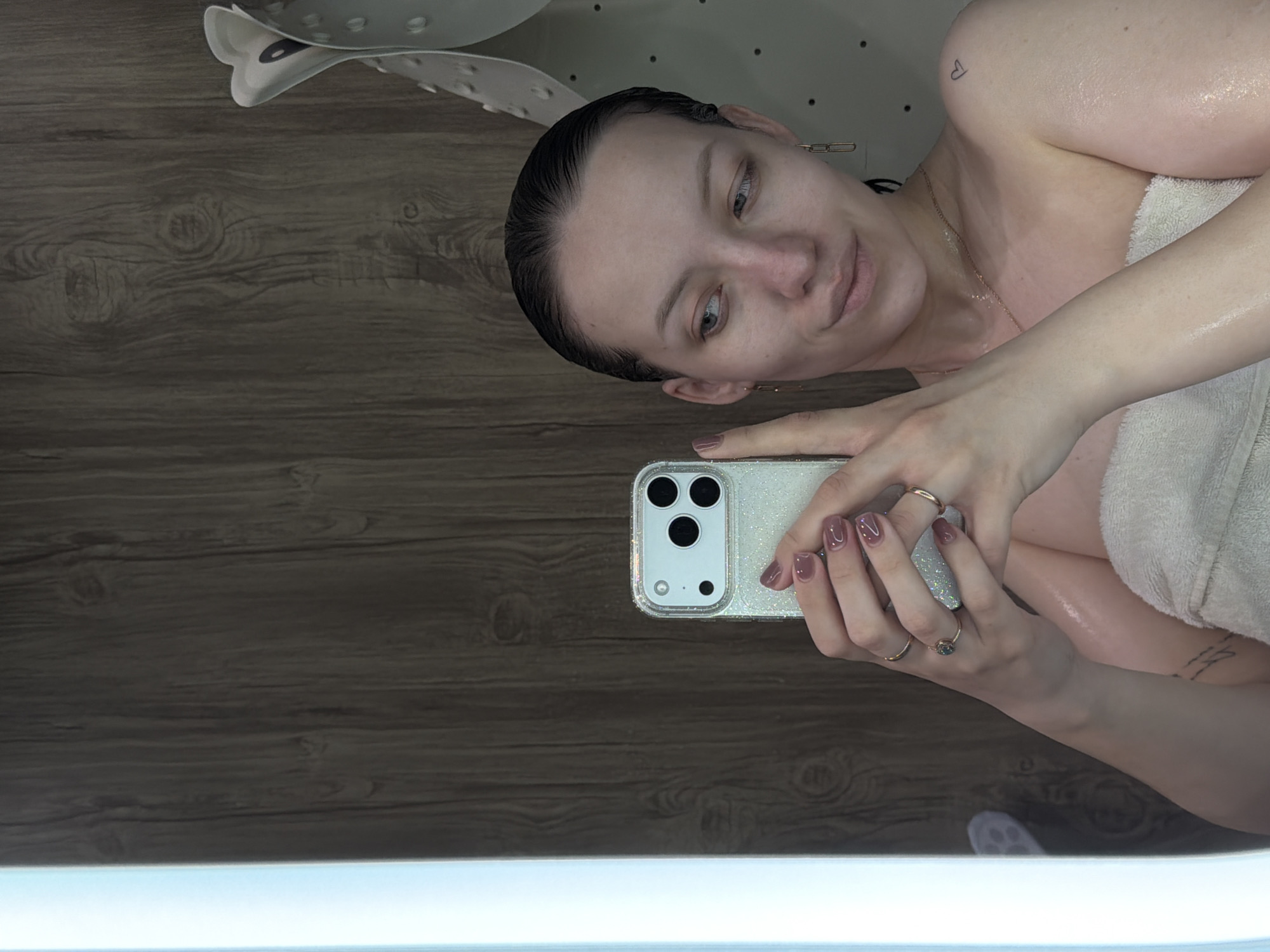 A woman is taking a selfie in a bathtub. She is fair-skinned with wet hair, wearing a gold chain necklace and wrapped in a white towel. She is holding a white smartphone with a clear glittery case. The bathroom has wood-look paneling on the walls. Part of a white bath mat is visible at the top. The location in the city of Reshetnikovo, Russia is not discernible from this image.