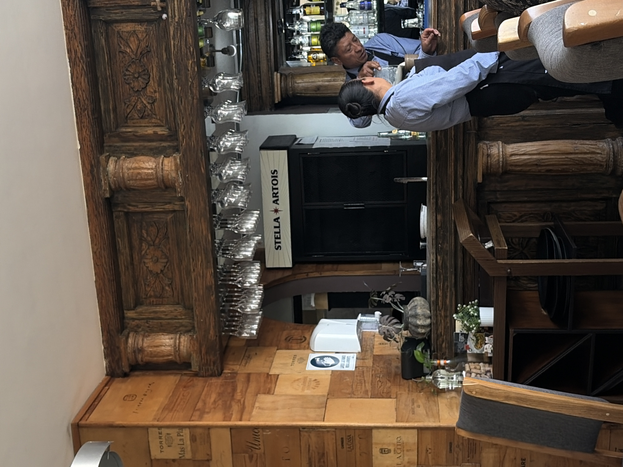 The image captures an interior scene in a restaurant in Puebla, Mexico. Two men, presumably employees, are at a bar. The bar's design includes a display of numerous wine glasses, a Stella Artois-branded cooler, and wooden elements with intricate carvings. The flooring is crafted from wooden crates repurposed as tiles. A hand-sanitizing station is also visible, as well as decor such as pumpkins and potted plants.
