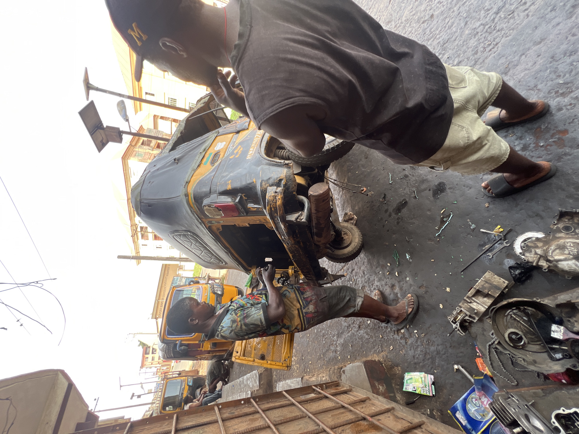 This image captures an active outdoor vehicle repair workshop in Nkpor, Nigeria, under bright daylight. The main subjects are two men engaged in repairing a yellow and black tricycle, commonly known as a "keke napep."
In the foreground, a man wearing a dark t-shirt, light-colored shorts, and a dark cap with a prominent "M" on it, stands leaning over the rear section of the tricycle. His hands appear to be working on a component near the back wheel. The other mechanic, visible in the mid-ground, lies on his back beneath the tricycle. He is dressed in a vibrant, multi-colored patterned shirt and shorts, actively working on the underside mechanics of the vehicle.
The setting is a somewhat dishevelled workshop floor, stained dark with oil and grime, and littered with various tools and vehicle parts. Scattered across the ground are engine blocks, spanners, wrenches, chains, and other components suggesting an ongoing engine or transmission repair. Notable parts include a disassembled engine case and what appears to be a cylinder head. In the background, other similar tricycles are parked, and buildings are visible under a bright, overcast or hazy sky.
Visible text on the main tricycle includes a registration number, "NRG0078." On some packaging on the ground, the letters "SAE" are discernible, likely indicating an oil specification. The overall scene conveys a busy, hands-on environment dedicated to vehicle maintenance and repair.