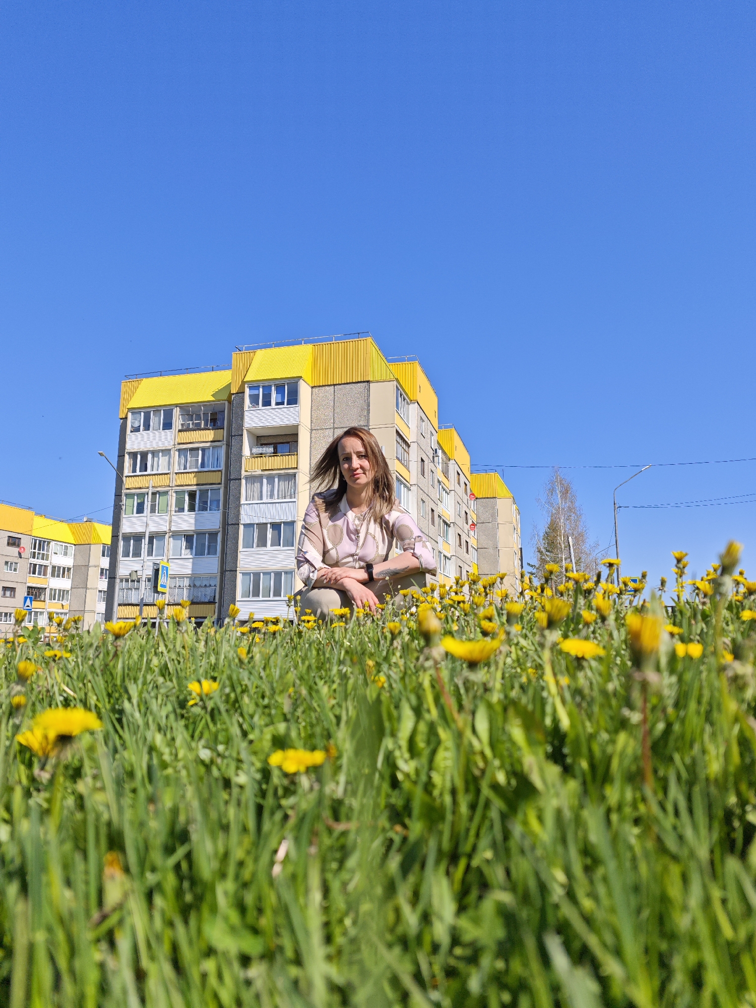 A medium-shot captures a woman crouching in a field of dandelions in Марковский, Russia, with a multi-story apartment building in the background under a clear blue sky. The apartment building features yellow trim above each floor. The woman wears a patterned shirt and light-colored pants. The dandelions are in full bloom, creating a vibrant foreground. A street lamp is visible on the right side.