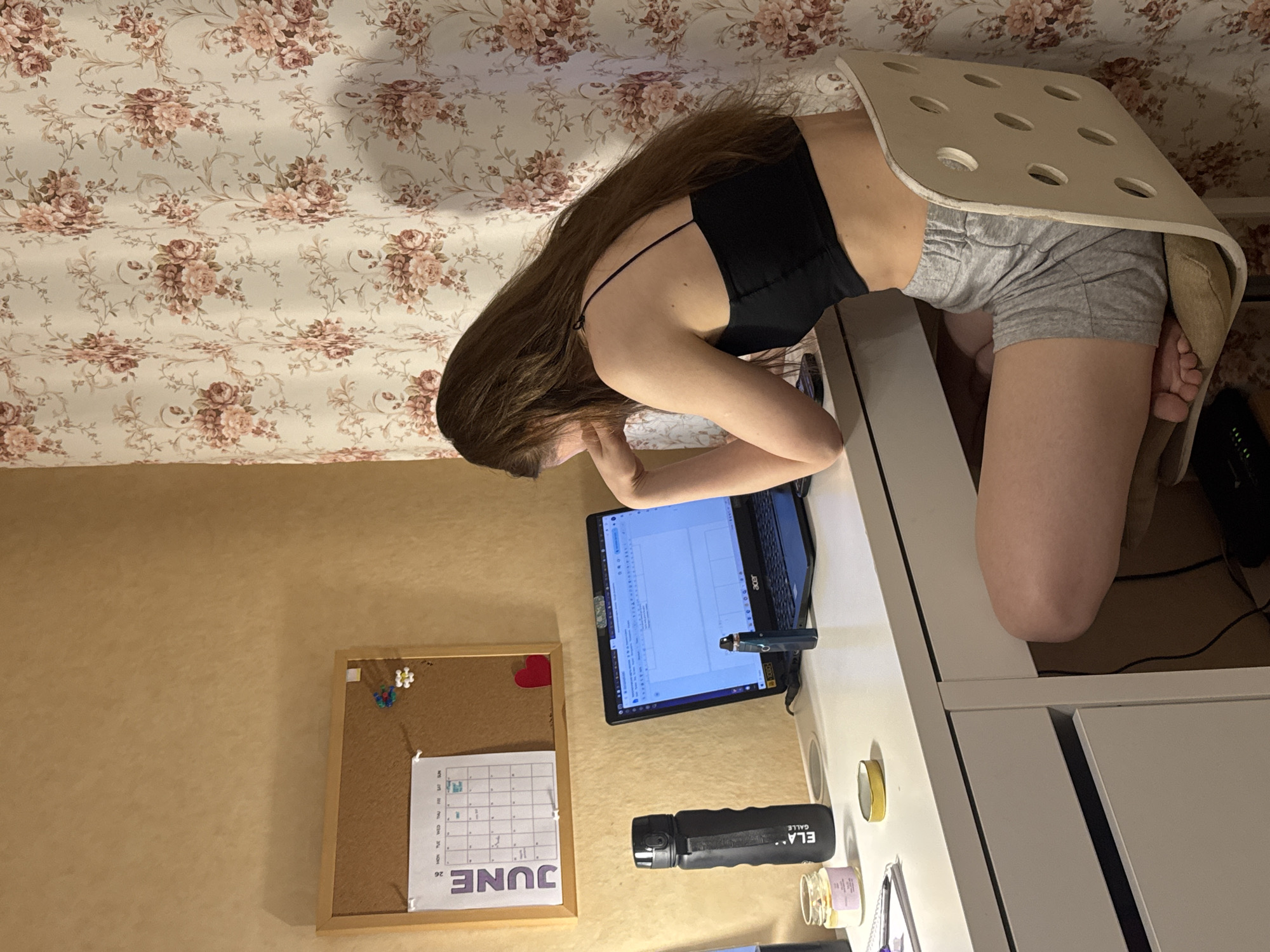 This indoor scene, situated in Perm, Russia, depicts a female individual seated at a white desk, concentrating on an open laptop. The individual has long, dark brown hair and is dressed in a black spaghetti strap crop top and grey athletic shorts. She is positioned on a light-colored wooden chair featuring a geometric pattern of circular cutouts on its backrest, with her bare feet tucked beneath the seat. Her left elbow rests on the desk, supporting her head as she gazes at the laptop screen.
The white desk is occupied by several objects, from left to right: a small glass jar containing a yellow substance, a black water bottle marked "ELA CALLE" vertically, a circular gold-colored lid, and a black vape pen standing upright next to the laptop. The laptop, an Acer model, is open, displaying what appears to be a document editing or spreadsheet application. A black power cable is visible connecting to the laptop from below the desk.
The wall behind the desk on the left is a textured, light beige color. A cork board with a wooden frame is mounted on this wall, featuring a "JUNE" calendar with the number "26" prominently displayed, alongside various colorful pushpins, including a flower shape, and a red heart-shaped object pinned below the calendar. To the right, a white fabric curtain with a repeating pattern of pink and rose-colored flowers and green foliage hangs, partially obscuring the background.