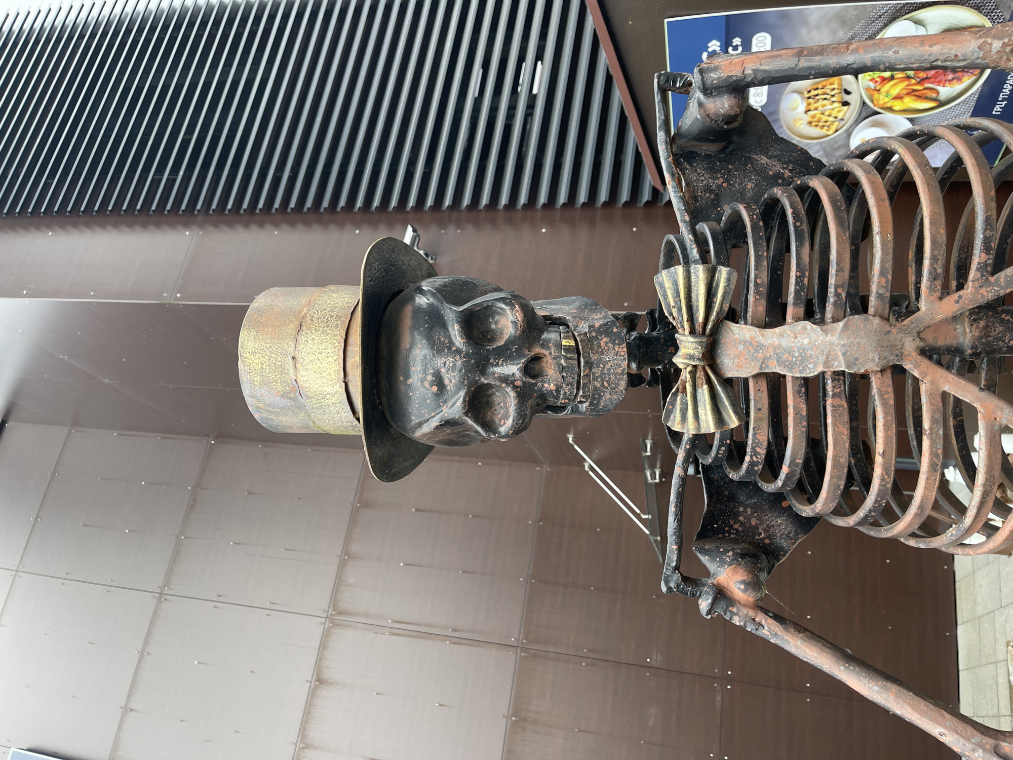 A metal sculpture of a skeleton wearing a top hat and a bowtie stands in front of a building with a textured, brown facade and a darker, ribbed section on the right. The skeleton is made of dark metal with rust-colored highlights. It has a visible rib cage, arms, and a skull with a grimacing expression and gold teeth. A poster with images of food is partially visible on the lower right. The background suggests an outdoor urban environment, possibly a commercial area in Zelenogradsk, Russia.