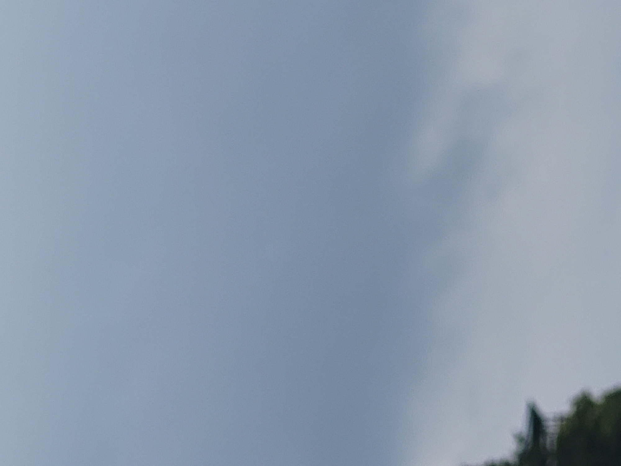 The image depicts a cloudy sky with a hint of green foliage and a dark, indistinct structure in the lower left corner. The structure appears to be a building with a roof, possibly a pavilion or a small house. Due to the low resolution and blurriness, specific details about its architecture or the surrounding environment are not discernible. The overall impression is that of an outdoor scene, likely captured during overcast weather. The location in Ozolnieki, Latvia, cannot be confirmed from the visual information present.