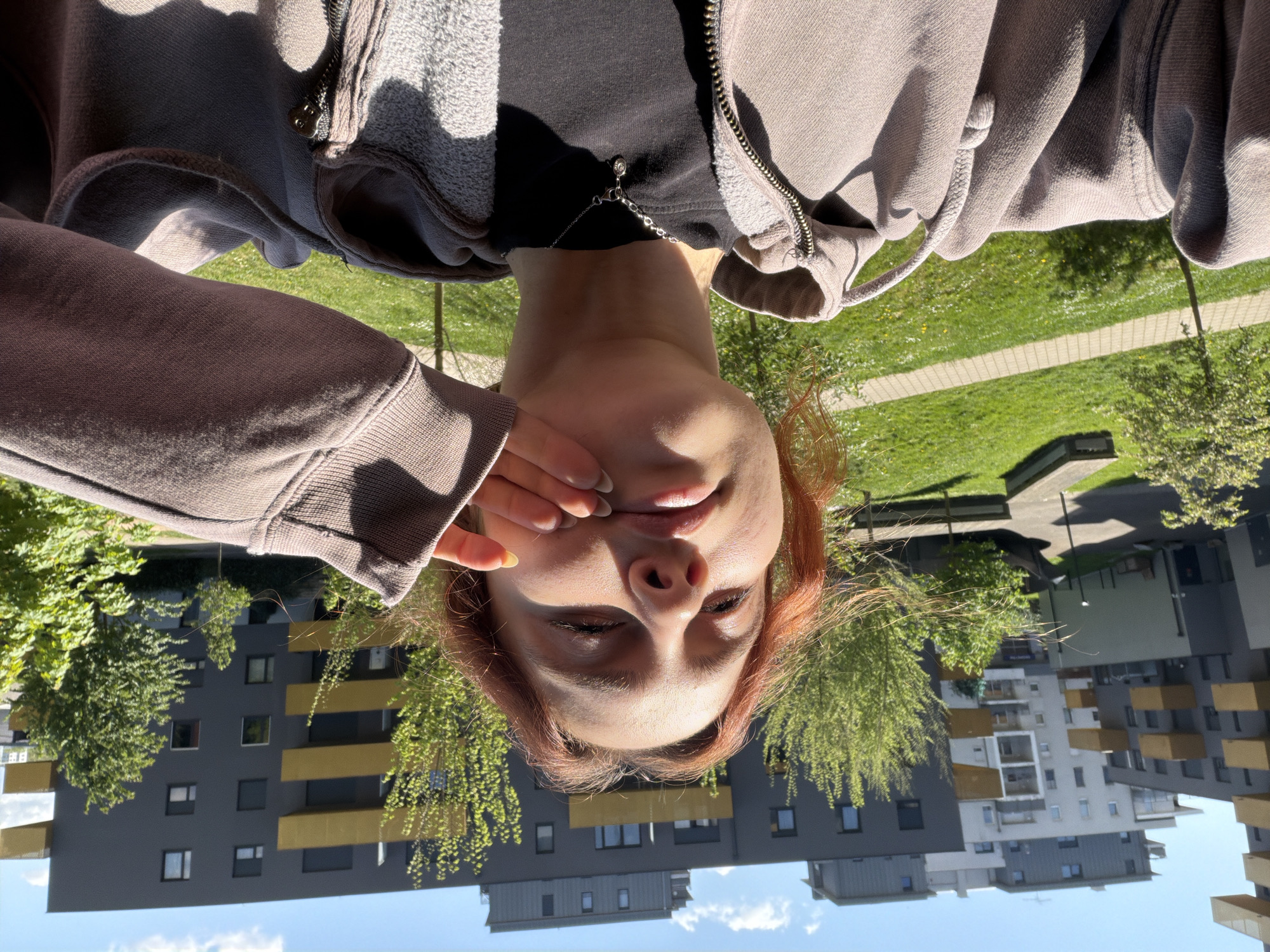 A woman with red hair is seen from a low angle, inverted perspective, against an outdoor background. She wears a zippered hoodie over a black shirt and a delicate chain necklace. Her hand is raised to her mouth. Behind her are modern buildings with a gray facade and gold-colored balconies, trees with fresh green leaves, a grassy area, and a blue sky with light clouds. Based on the architectural style and color scheme, the buildings are likely located in the Rivetoile district of Strasbourg, France.