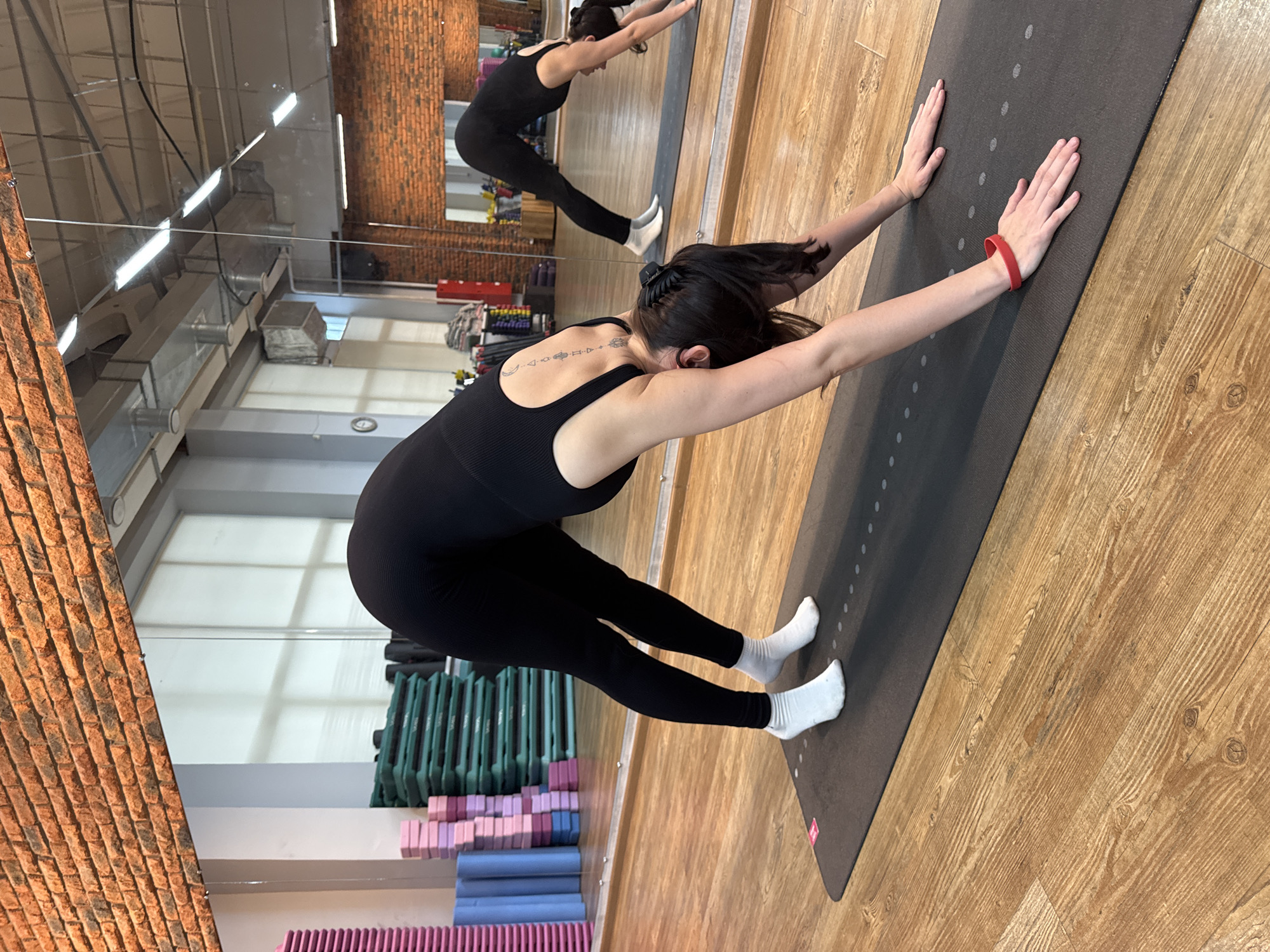 In a yoga studio in Chaikovsky, Russia, a woman with dark hair, wearing a black yoga bodysuit and white socks, is performing a yoga pose. She is facing away from the camera, with her arms extended forward and her palms flat on a dark yoga mat. Her body is in a forward fold, with her hips pushed back and her torso angled towards the floor. She has a red fitness tracker on her left wrist. In the background, a mirror reflects another woman in a similar pose, and stacks of colorful yoga blocks and mats. The studio has wooden floors and exposed brick walls, suggesting a modern and functional space. The lighting appears to be artificial, consistent with an indoor environment. The overall atmosphere is one of focused physical activity and calm.