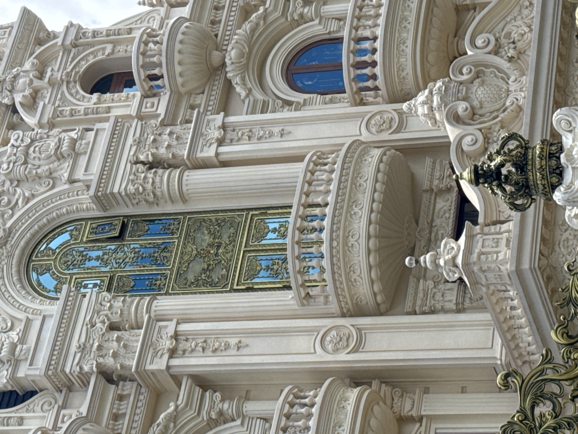 The image shows a close-up of the facade of a large, ornate building with classical architectural elements. The building is constructed of light-colored stone and features elaborate carvings, columns, balconies with balustrades, and arched windows. One large window, possibly a door, has intricate golden metalwork and blue glass. Another arched window with a wooden frame and blue glass is visible on a smaller balcony. The style suggests a grand, possibly historical or very elaborate modern structure. The overall impression is one of opulence and detailed craftsmanship. This building is identified as the Palace of Da Lat in the city of Da Lat, Vietnam.