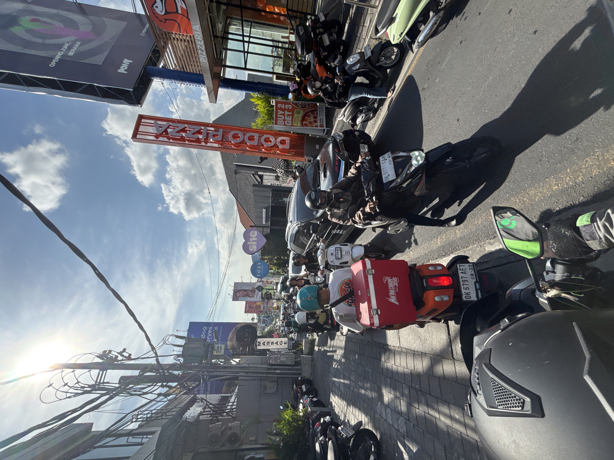 The image shows a busy street scene in North Kuta, Indonesia. A long line of motorcycles is stopped in traffic, with riders visible on some. Several businesses and signs are visible along the street, including "DODO PIZZA" and various advertisements for beauty and wellness services ("MASSAGE", "she", "AMY", "FOXY"). A sign indicates the opening of a new business named "PONY" in June, located in Berawa. The sky is partly cloudy, with the sun shining brightly. Power lines are strung overhead. A license plate on one of the motorcycles reads "DK 6797 AET", and another reads "DK 5171 CAH". One motorcycle has a delivery box for a company called "Marina comen 355".
