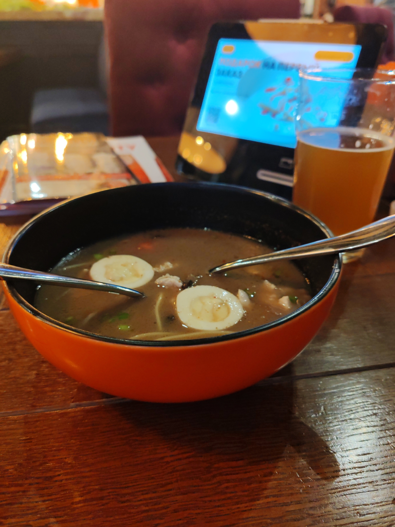 The image captures an indoor dining scene, likely in a restaurant or cafe in Saint Petersburg, Russia, during the evening. In the foreground, a vibrant orange and black bowl holds a hearty soup, resembling ramen or a similar noodle dish. The soup features a dark broth, visible noodles, two neatly halved hard-boiled eggs with pale yolks, small pieces of meat (possibly chicken or pork), and scattered green garnishes. Two silver spoons are positioned within the bowl.
To the right of the bowl, a glass contains an amber-colored liquid, consistent with beer. The items rest on a dark wooden table with a visible grain. In the midground, a tablet device is prominently displayed, its screen glowing bright blue. Partially visible Russian text on the screen reads "ПОДАРОК НА ПЕРВЫЙ" (GIFT FOR THE FIRST), suggesting a promotional offer or part of a digital menu/ordering system. To the left, a blurry object, possibly a plastic-wrapped menu or another food item, is seen. The background is softly blurred, showing hints of dark red or purple upholstered seating, confirming an indoor setting with low, ambient lighting, indicating nighttime or a cozy atmosphere. The scene depicts a relaxed meal in progress or about to commence.