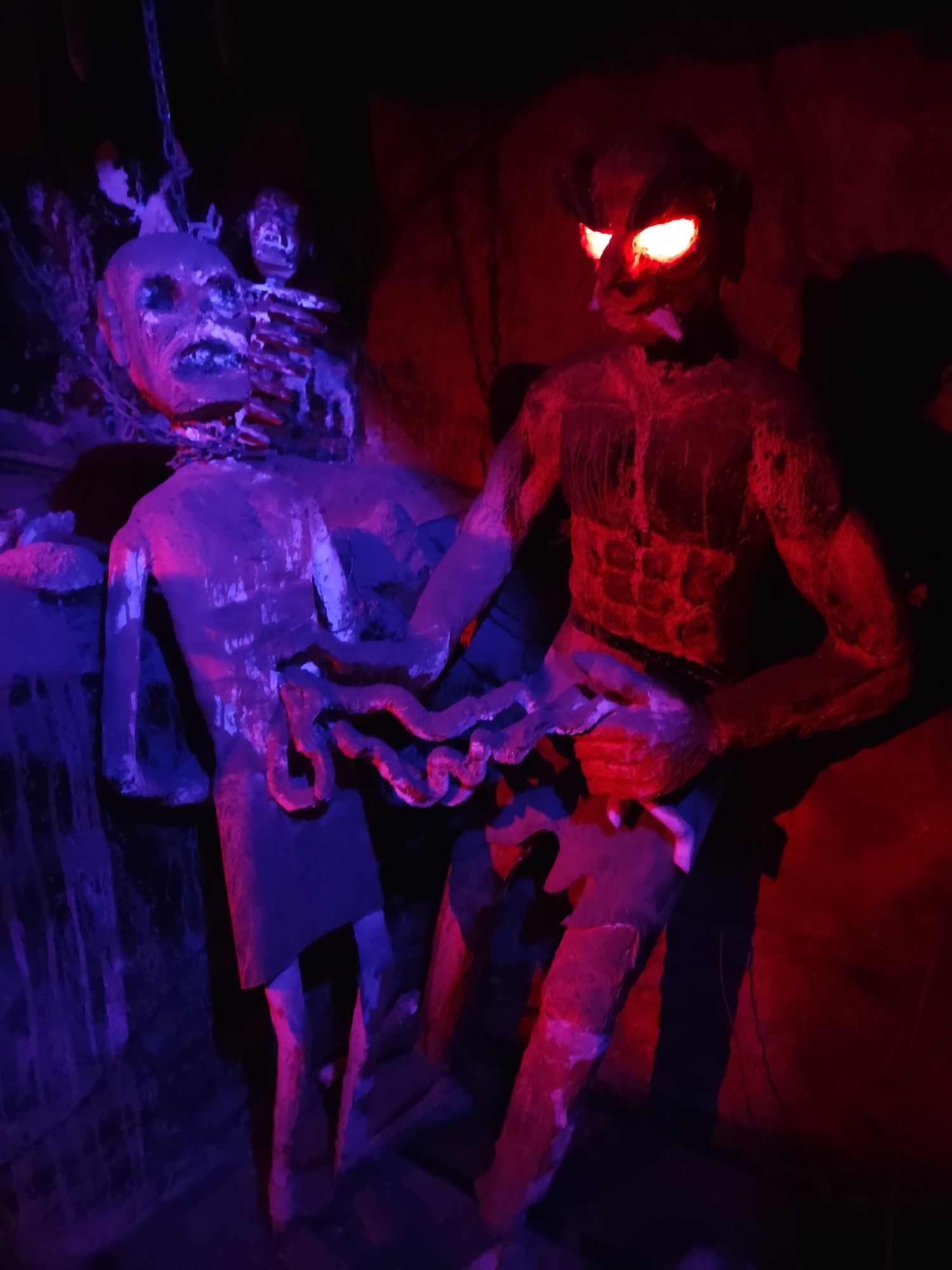 The image depicts a macabre scene with two sculpted figures illuminated by red and purple lights. On the left, a figure with a disfigured face and exposed rib cage is chained by the neck. Its body is painted in shades of purple and blue, with visible chains and a skeletal structure. To the right, a muscular, demonic-looking figure with glowing red eyes and horns is positioned, its body painted red. This figure appears to be interacting with the chained figure, holding what looks like intestines. The background is dark and rocky, suggesting a cave-like or dungeon setting. The lighting creates a dramatic and unsettling atmosphere.