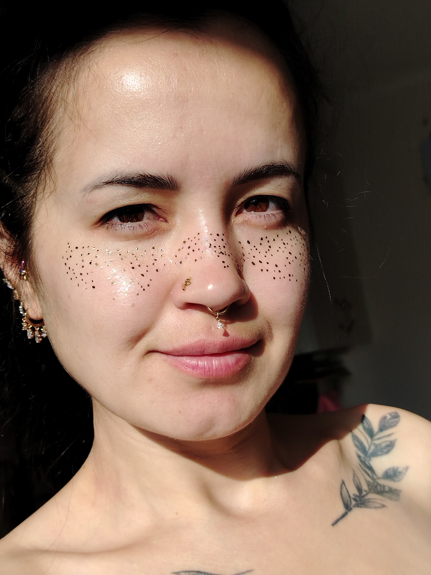 A close-up, portrait shot shows a young woman from the chest up, facing the camera with her head slightly turned to her left. Her skin is illuminated by direct sunlight, creating a highlight across her forehead and cheek. She has dark hair, pulled back from her face, and is wearing several pieces of jewelry: a small hoop earring with multiple dangling charms on her left ear, and a gold septum ring with a small white gem. Her face is adorned with numerous small, gold glitter dots, mimicking freckles, concentrated across her cheeks and the bridge of her nose. Her lips are lightly colored, and she has a subtle smile. A tattoo of a branch with leaves is visible on her right shoulder. The background is dark and out of focus, with hints of a room and possibly a window to the right. The scene is indoor, likely in a residential setting. The image is a still photograph and does not depict any action or interaction within Noviy, Russia.