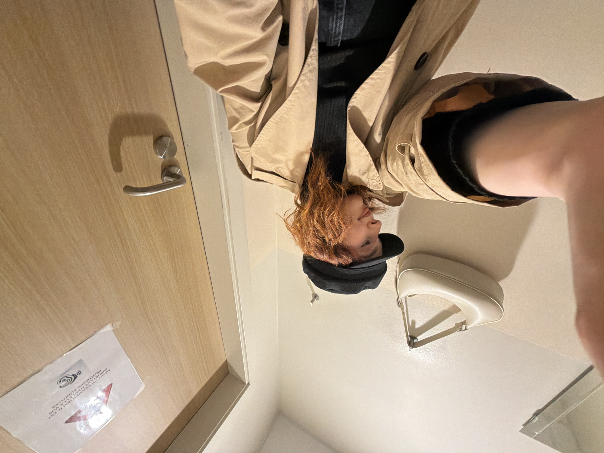 A person wearing a black cap, tan trench coat, and black top is standing in what appears to be a bathroom or dressing area. A white purse is hanging on the wall. The scene includes a door with a silver handle and a white sign with red and black text. The location cannot be identified as Strasbourg, France.