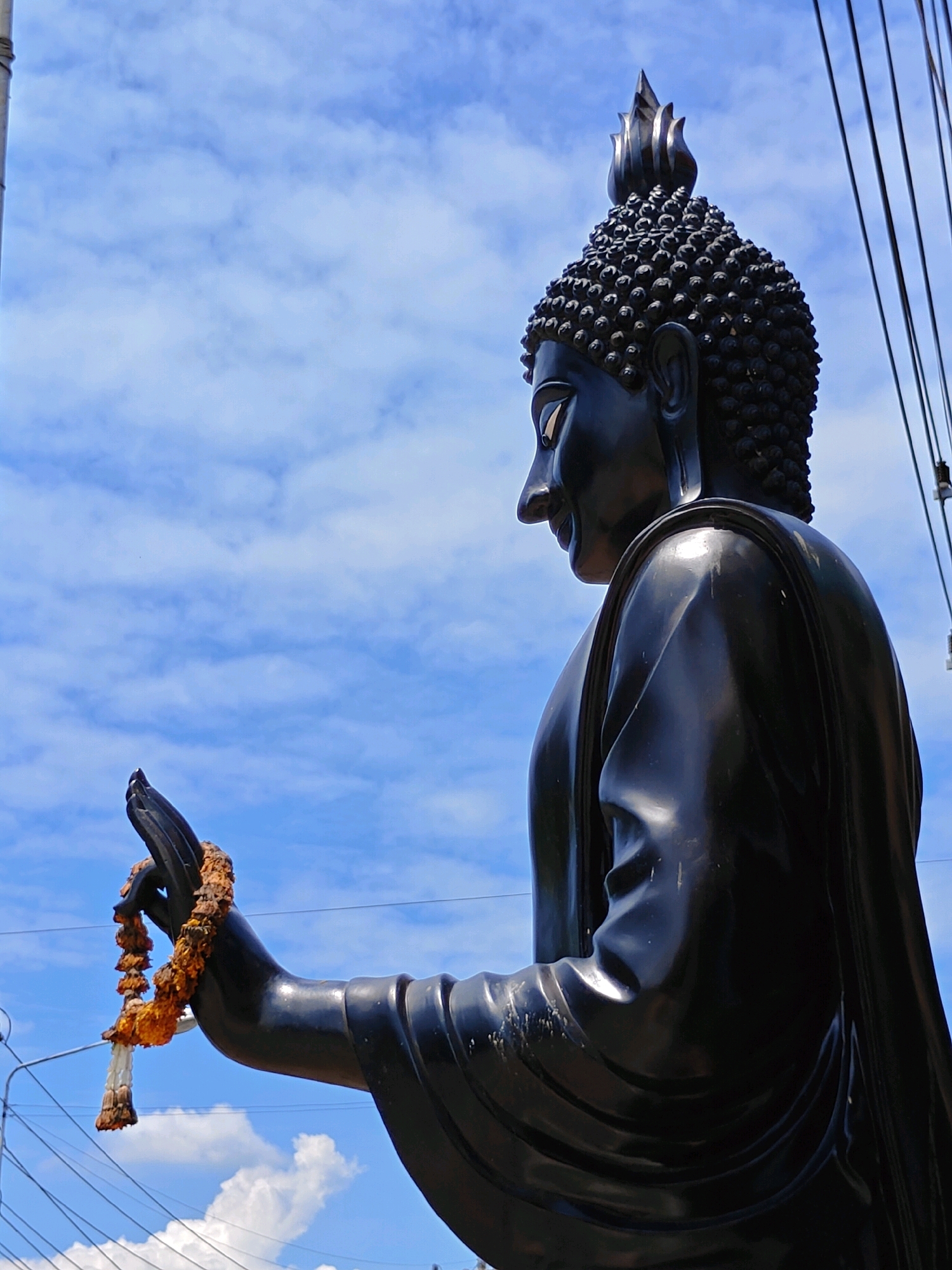 A full-body, side-profile, medium-shot photograph captures a large, dark, metallic Buddha statue against a bright blue sky dotted with white clouds. The statue is depicted from the waist up, with its head turned slightly to the left and its right hand extended forward, holding a wilting garland of orange and white flowers. The Buddha statue wears a traditional robe, which drapes over its shoulders and torso, with visible creases and folds. The statue's face shows a serene expression, with closed eyes and a slight smile. Electrical wires are visible diagonally across the sky on the right side of the frame. The overall scene suggests an outdoor setting, likely in a public space within the city of Phra Nakhon Si Ayutthaya City Municipality, Thailand, possibly a temple or a roadside shrine, given the presence of the statue and the overhead wires.