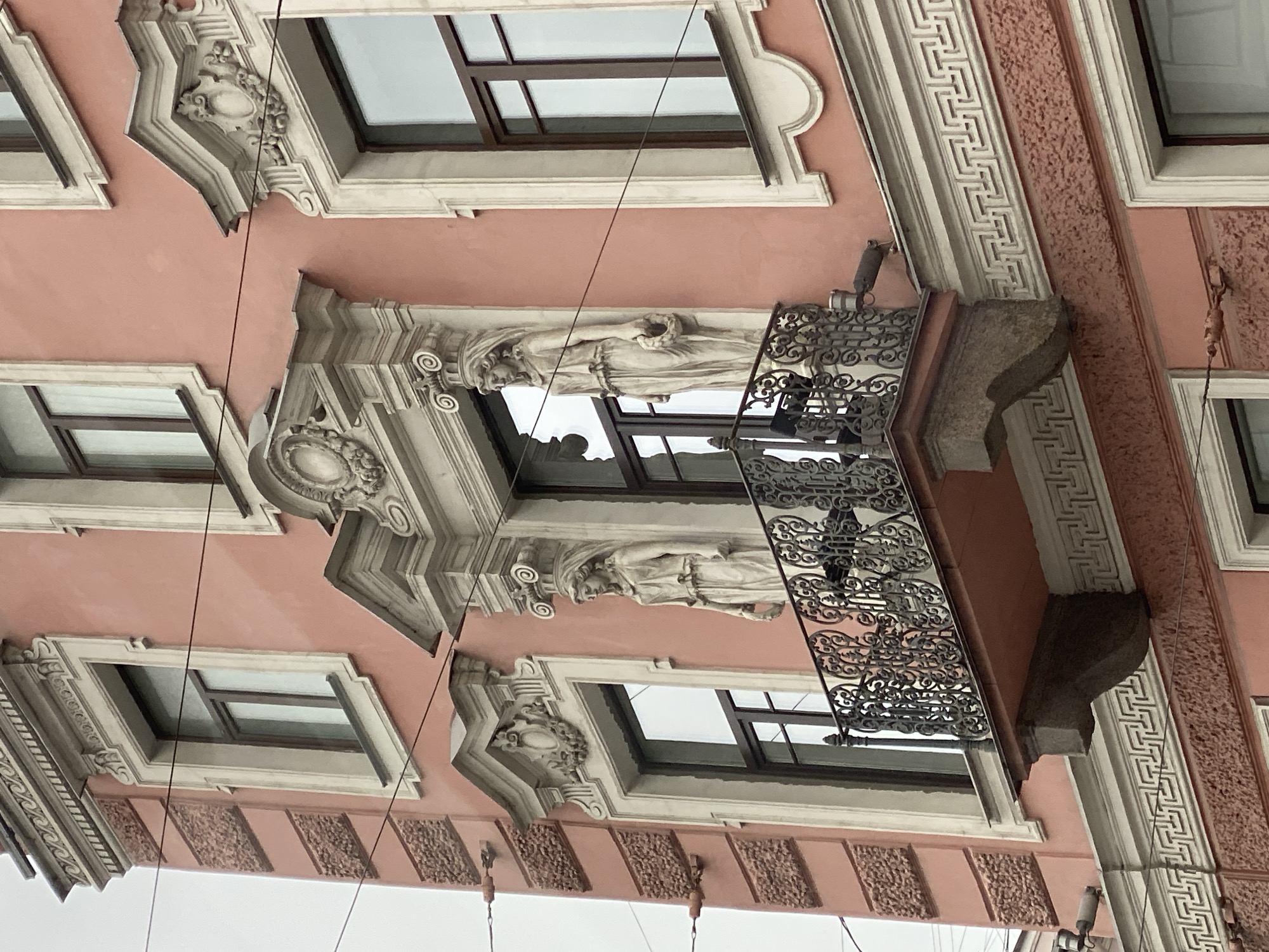 A low-angle shot depicts the ornate facade of a building with multiple windows and decorative elements. The building has a pale pink exterior. The windows have dark frames and are recessed within the facade, surrounded by white, decorative moldings. A balcony with a wrought-iron railing extends from the building, featuring complex and artistic designs. Above the balcony, there are sculptures of human figures, positioned near the windows, contributing to the building's architectural embellishment. The upper part of the building is visible, with its cornice and additional decorative elements. The sky, visible through the windows, is overcast and pale. There are utility wires running across the foreground of the image.