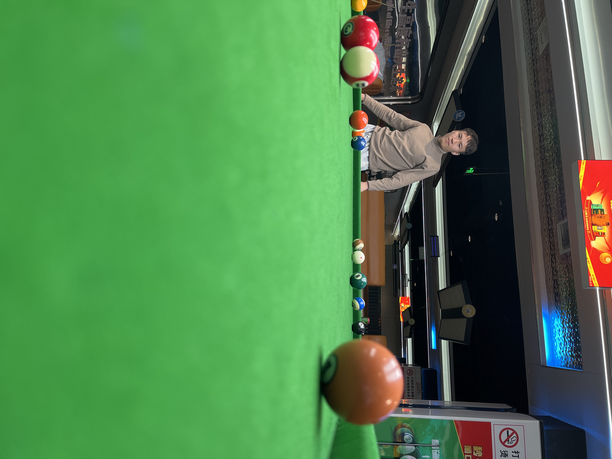 The image depicts a person playing billiards in an indoor entertainment venue, possibly a billiard hall in Daxing District, China. The scene is dominated by the vibrant green felt of a pool table, with several billiard balls arranged in a line. The person, a young male dressed in a casual beige long-sleeved shirt, is leaning over the table, appearing focused on the game. Various billiard balls, some solid colors and others with stripes, are visible, including a prominent orange cue ball in the foreground.
In the background, the setting suggests a modern establishment with reflective surfaces, ambient lighting, and possibly other gaming areas or seating. The large windows in the background offer a glimpse of the outside, though the time of day is not clearly discernible. Signs with Chinese text are visible, including one that warns against smoking while playing billiards to prevent damage to the table. The overall atmosphere appears relaxed and recreational, with the individual engrossed in the game.