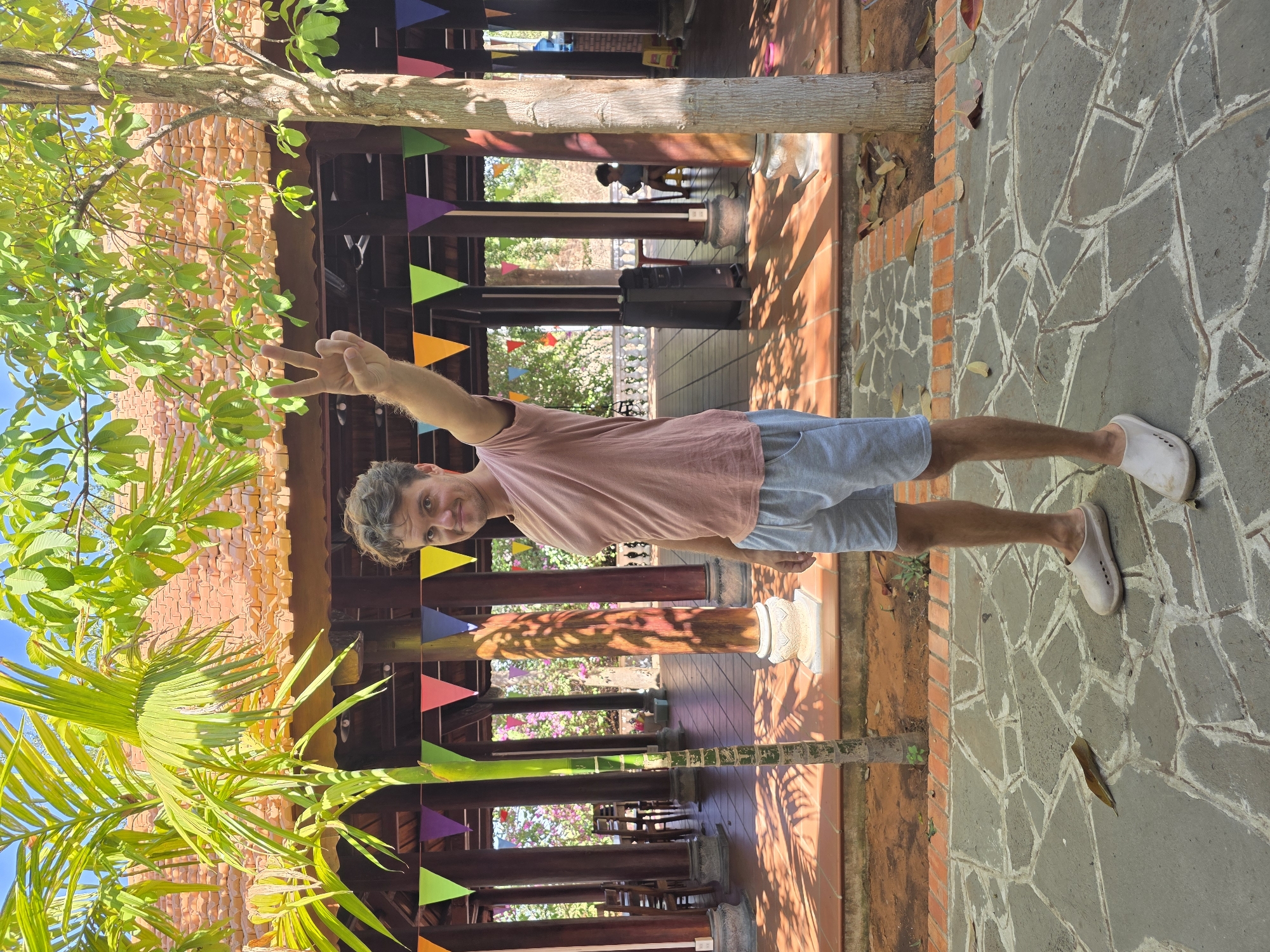 A man stands on a stone walkway in Mũi Né, Vietnam, facing the camera and giving a peace sign with his right hand. He wears a pink t-shirt, blue shorts, and white clogs. The walkway is made of irregularly shaped gray stones with white grout and is bordered by orange brick. Behind the man is a building with a red tile roof and wooden pillars decorated with colorful pennants. A lush green palm tree is visible on the left side of the frame, and another tree is situated behind the man. In the distance, through the building's open structure, are more trees, buildings, and a person sitting on a chair.