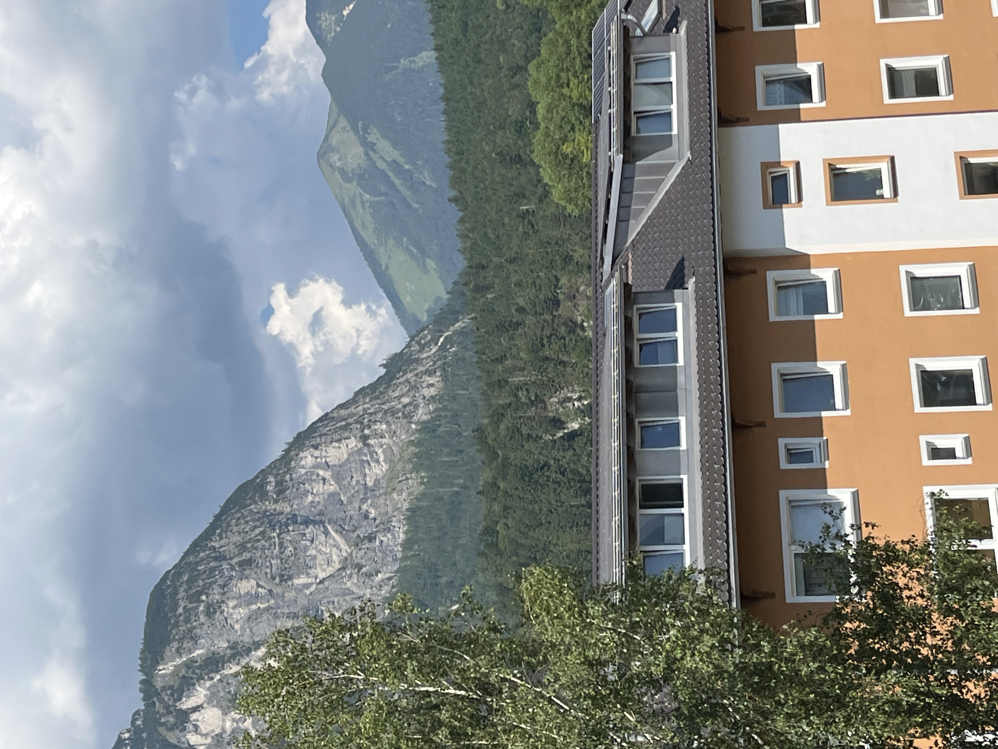 The image shows a multi-story building with orange and white facades, featuring numerous windows. The building's roof is dark and angled, with some sections covered in solar panels. In the background, a dense forest covers the lower slopes of steep, rugged mountains. The sky above is partly cloudy, with dramatic formations of white and dark gray clouds. A tall, leafy tree is visible on the left side of the frame, partially obscuring the building and the mountainous landscape. The scene appears to be a view from a higher elevation looking down towards the building and across to the mountains, likely taken from another building or a vantage point within the city of Garmisch-Partenkirchen, Germany.