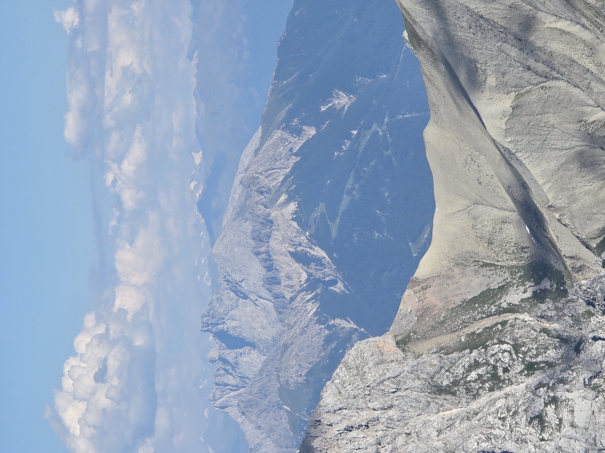 The image depicts a panoramic view of mountainous terrain, likely in or near Garmisch-Partenkirchen, Germany, given its characteristic alpine landscape. The foreground features a steep, rocky slope covered with sparse patches of green vegetation, indicative of high-altitude scree and alpine meadows. This rugged section of the mountain is rendered in shades of gray and brown, with sharp textural details from the rocks and the shadows cast upon them.
Moving into the midground, the landscape transitions to softer, more rounded mountain slopes covered in a dense, dark green forest. The contrast between the bare rock of the foreground and the forested slopes of the midground is stark. Further in the distance, a range of majestic, jagged mountains rises, their peaks appearing light gray and rocky, some possibly capped with snow or glaciers. These distant mountains exhibit a layered appearance, with multiple ridges and valleys creating a sense of depth and scale.
The sky above is a clear, bright blue, punctuated by scattered white, fluffy clouds. These clouds appear to be cumulus formations, common in fair weather conditions at high altitudes. The overall lighting suggests a sunny day, likely during the daytime, with the sun positioned to cast distinct shadows that highlight the rugged topography of the mountains.
The scene is devoid of any human presence or man-made structures, emphasizing the raw, natural beauty of the alpine environment. The composition of the image, with its layers of mountains receding into the distance, creates a feeling of vastness and grandeur. The forensic detail lies in the precise rendering of geological features: the erosion patterns on the rock faces, the texture of the forest canopy, and the distinct geological formations of the mountain ranges.