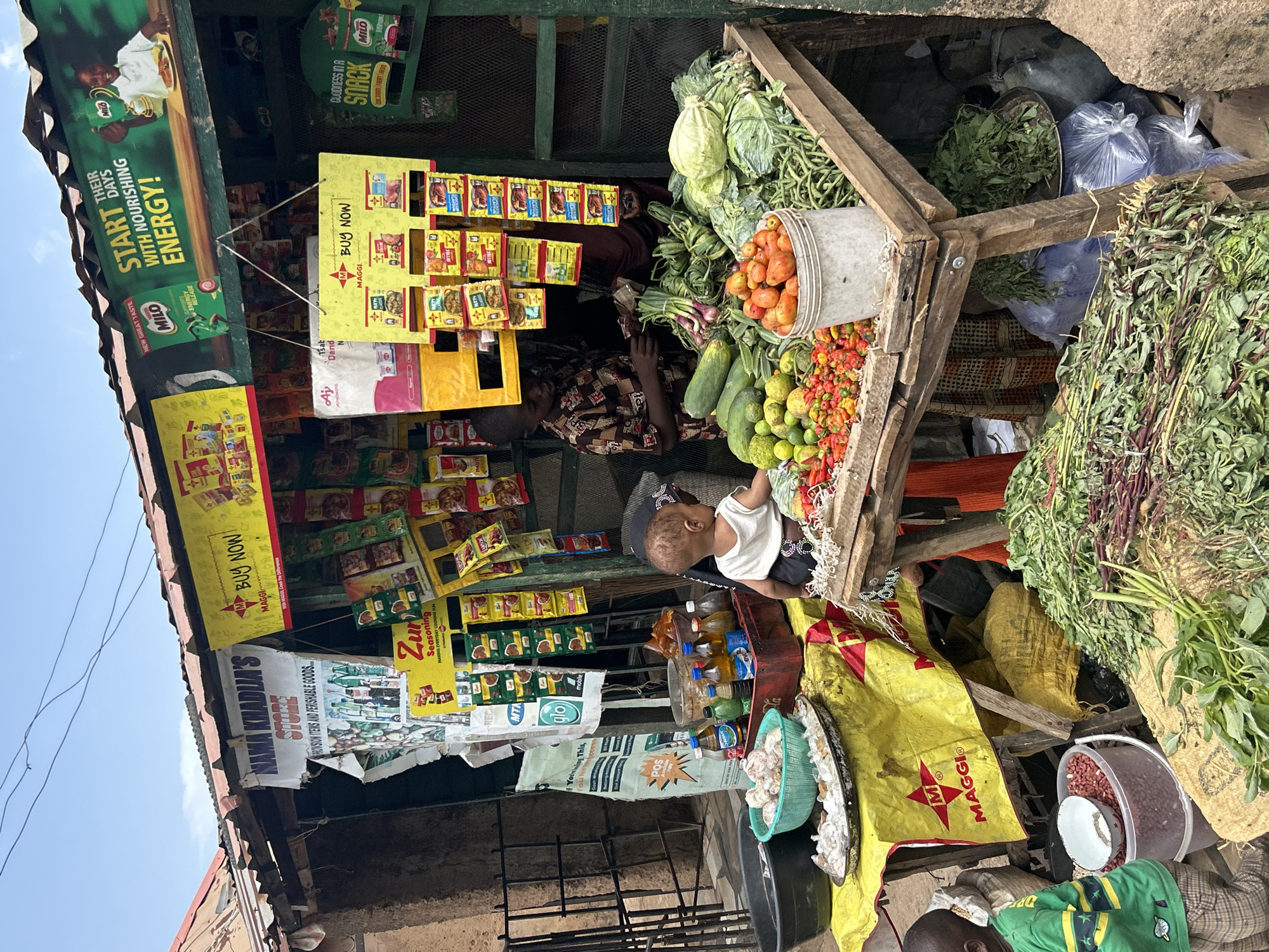 The image depicts a bustling roadside market stall in Jos, Nigeria. The stall, named "NAMA KHADIA'S STORE," is stocked with a wide variety of goods, including Maggi seasoning packets, Milo products, vegetables (cabbage, tomatoes, cucumbers, peppers), leafy greens, and bottled beverages. A young child is perched on the edge of the display, near the beverages. An adult is behind the vegetable display. The stall has promotional signs and advertisements. Bags of goods and other items are on the ground. The sky is visible in the upper-left corner.