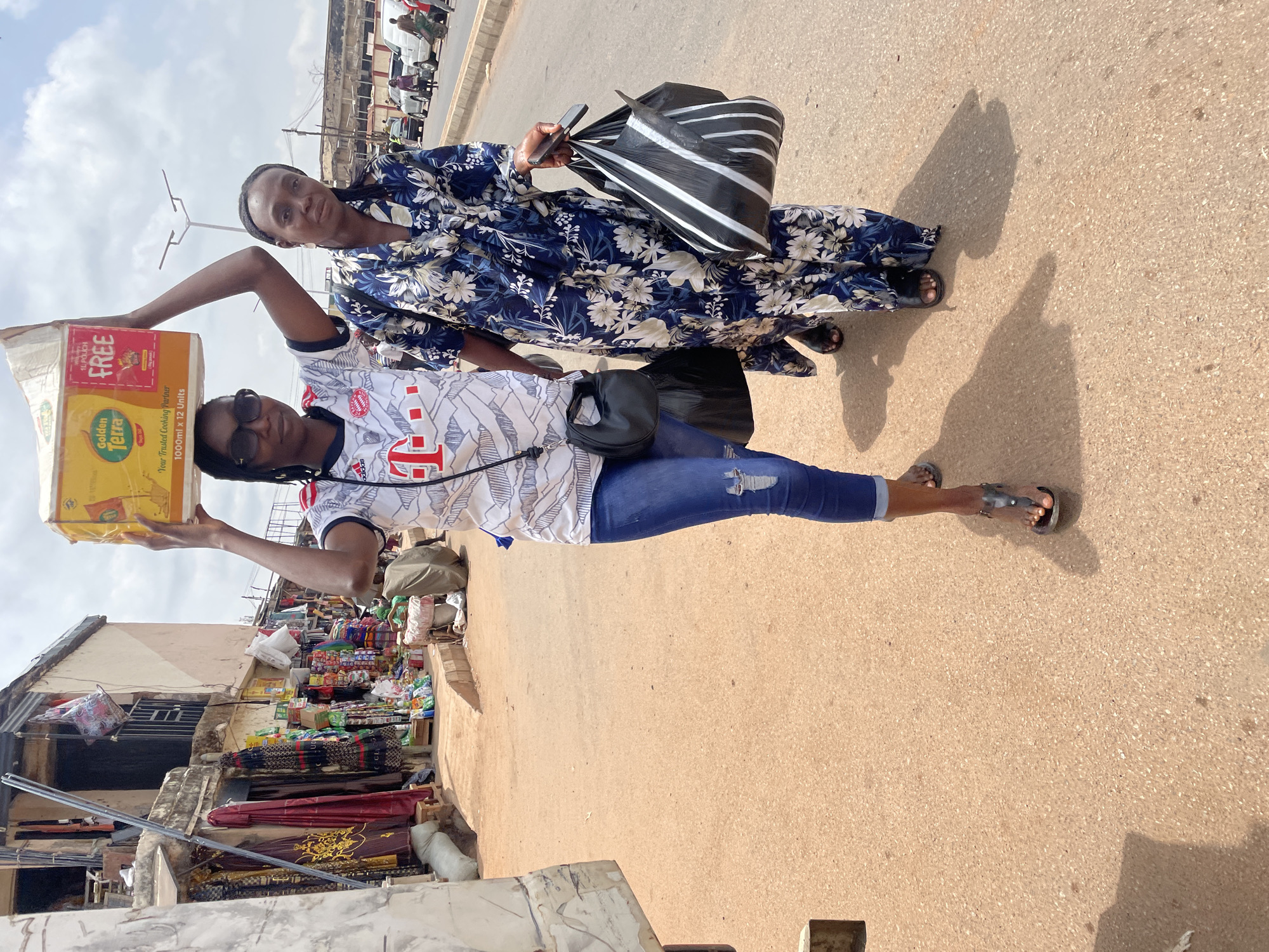 Two women stand on a paved road in Jos, Nigeria. The woman in the foreground balances a box of Golden Terra cooking oil on her head. She is wearing a white soccer jersey, blue jeans with a rip in the knee, sunglasses, and sandals. The other woman stands behind her, wearing a blue floral dress and sandals. She holds a black and white striped bag and a phone. Behind them is a street market with various goods displayed. The sky is bright with scattered clouds.