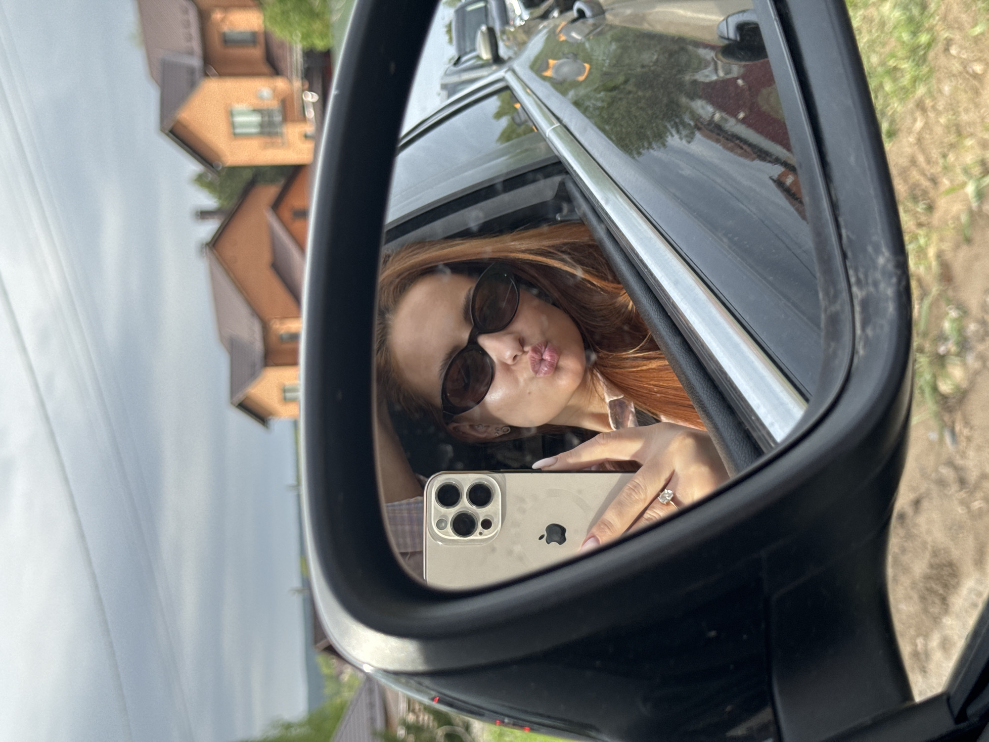 A woman with red hair and sunglasses is visible in the side mirror of a car. She is holding a smartphone, likely taking a selfie. The background seen through the car windows and in the mirror shows houses with reddish-brown roofs and green trees, indicating a residential area. The location is Гавриловка, Russia.