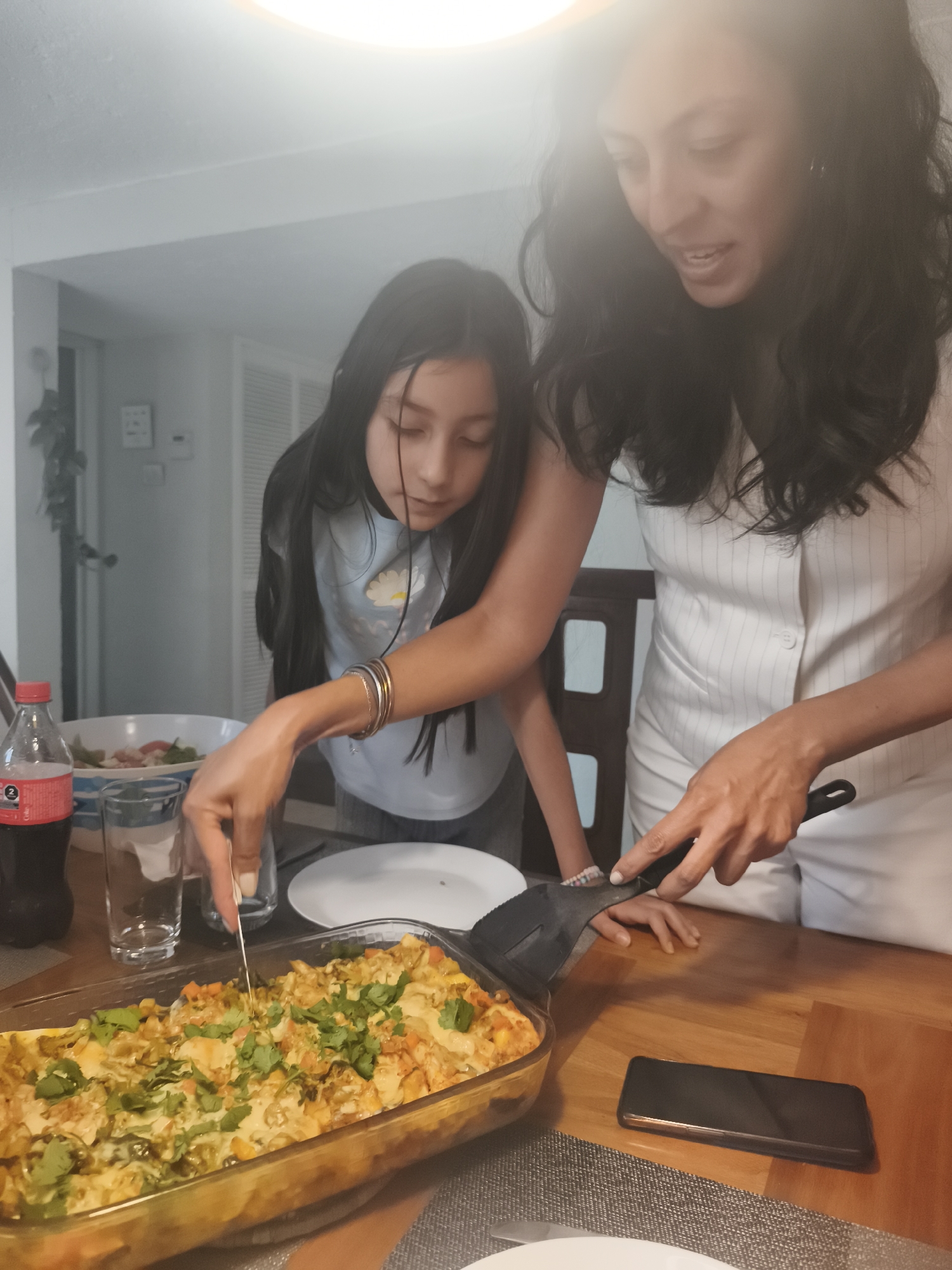 A woman and a young girl are cutting a casserole dish with a fork and spatula on a wooden table indoors. The casserole is in a glass baking dish and topped with herbs. A bottle of Coca-Cola, a glass, and a bowl of salad are also on the table. A cell phone rests on the table next to the casserole dish. The specific location is not discernible from the image, so it is not possible to determine if it is in Mexico City, Mexico.