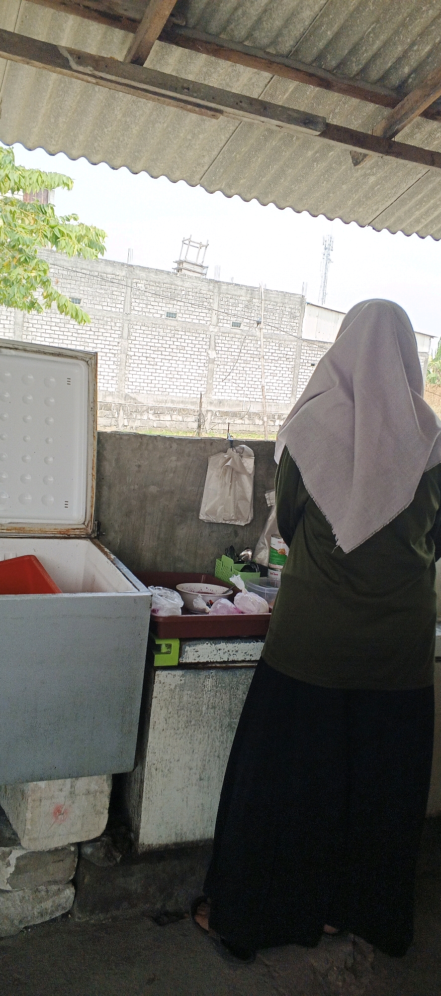 The image depicts an outdoor scene in Sonoadi, Indonesia, under a corrugated roof supported by wooden beams. In the foreground, to the left, is an open, light blue chest freezer with its lid ajar. Inside the freezer, a red plastic container is visible. To the right, a person wearing a light purple hijab and an olive green long-sleeved shirt over a black skirt stands facing away from the camera, toward a concrete counter. On the counter, several items are arranged, including a red tray with bowls, plastic-wrapped items, and a green plastic container holding utensils. Behind the counter, a plain concrete wall is visible, with a white plastic bag hanging from it. Beyond the wall, a brick building with a metal roof and a tall cellular tower are in the background, under a hazy sky. The ground is composed of dark, uneven paving stones or dirt. The scene appears to be an informal outdoor food stall or preparation area.