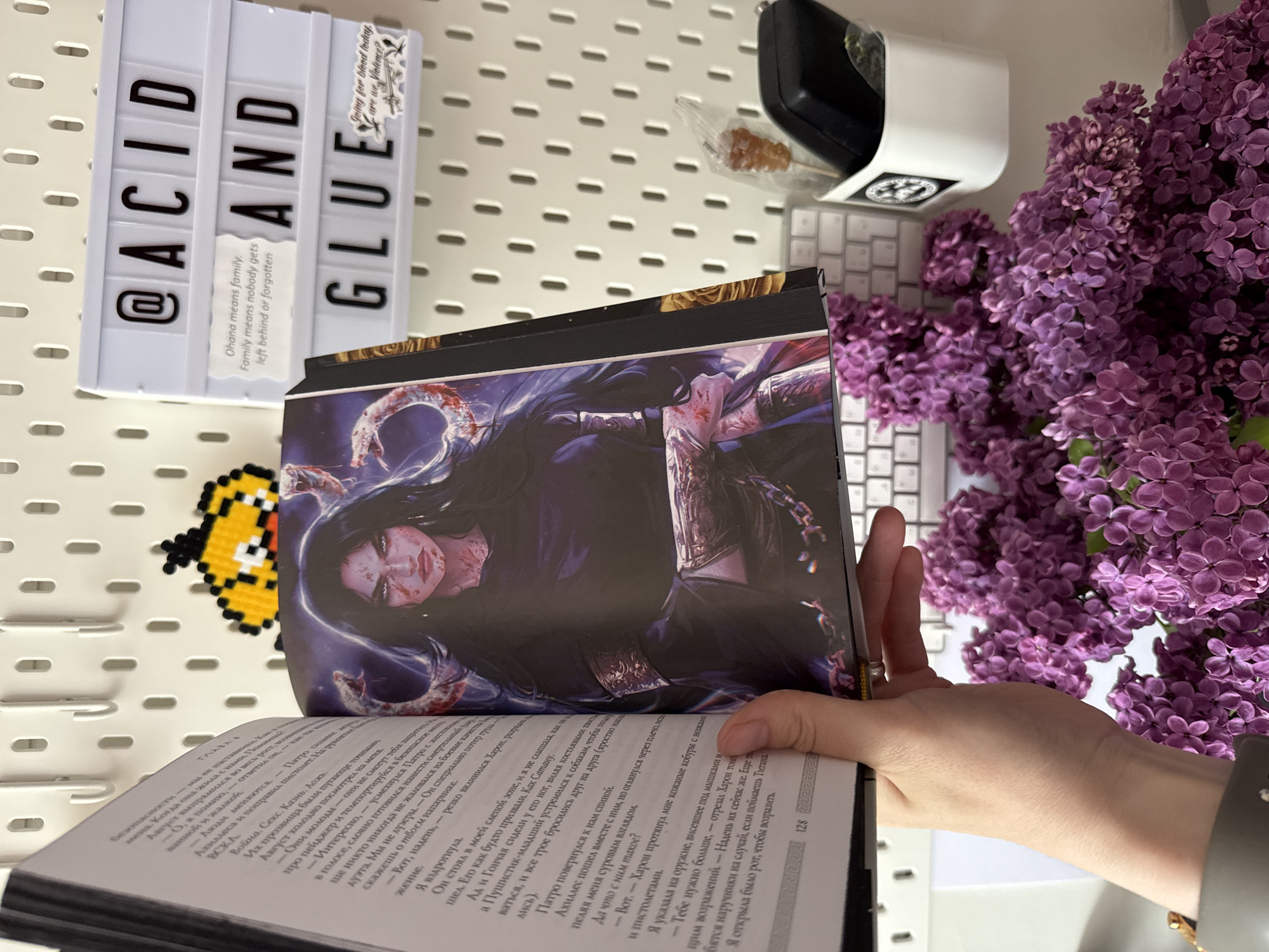 A book is open, held by a hand with a silver bracelet, revealing a color illustration on the right page and a Cyrillic text on the left. The illustration depicts a dark-haired, pale-skinned character with blood on their face and snakes around their head, against a dark background. The open book displays text in Cyrillic, with page number "128" visible at the bottom right. The text appears to be part of a novel.
Behind the book, there is a white wireless keyboard and a bunch of purple lilac flowers. To the left of the book, there is a white pegboard. On the pegboard are objects: a light box sign that reads "@ACID AND GLUE," with a small sticker attached to the right of the sign. To the right of the sign is an object wrapped in plastic. There is also a pixelated yellow Minion image hanging on the pegboard.
The scene gives the impression of being a personal workspace or desk. There is no indication of the location being Riga, Latvia.