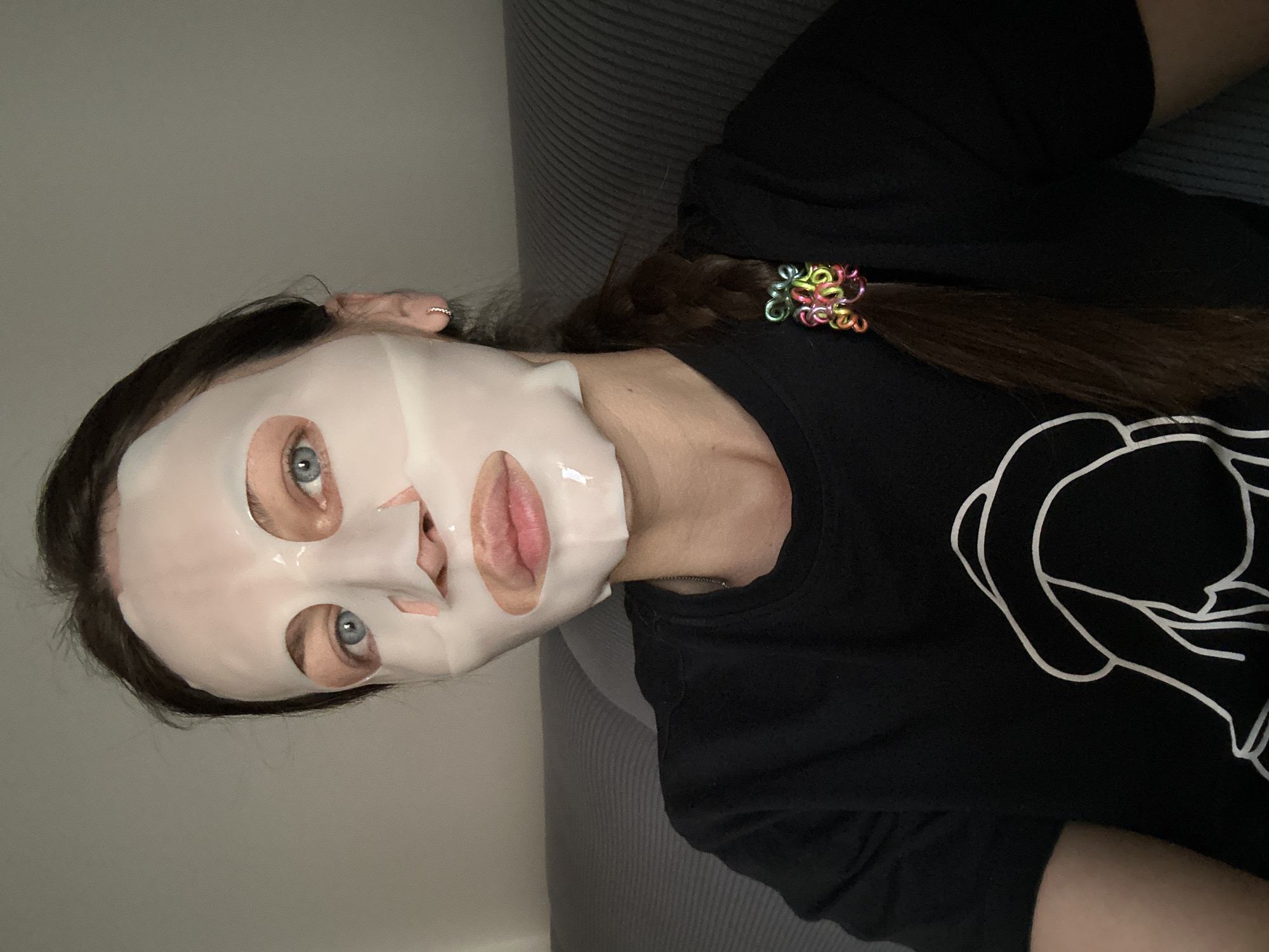 A person lies on a gray couch or sofa. They are wearing a black T-shirt with a white design on the front. The person has fair skin and blue eyes. They also wear a white facial mask, and their brown hair is pulled back in a braid with a colorful hair tie. A small ring is visible in the person's ear. The setting appears to be indoors with the person on a couch. The location in Donji Pobori, Montenegro, cannot be confirmed from the image.