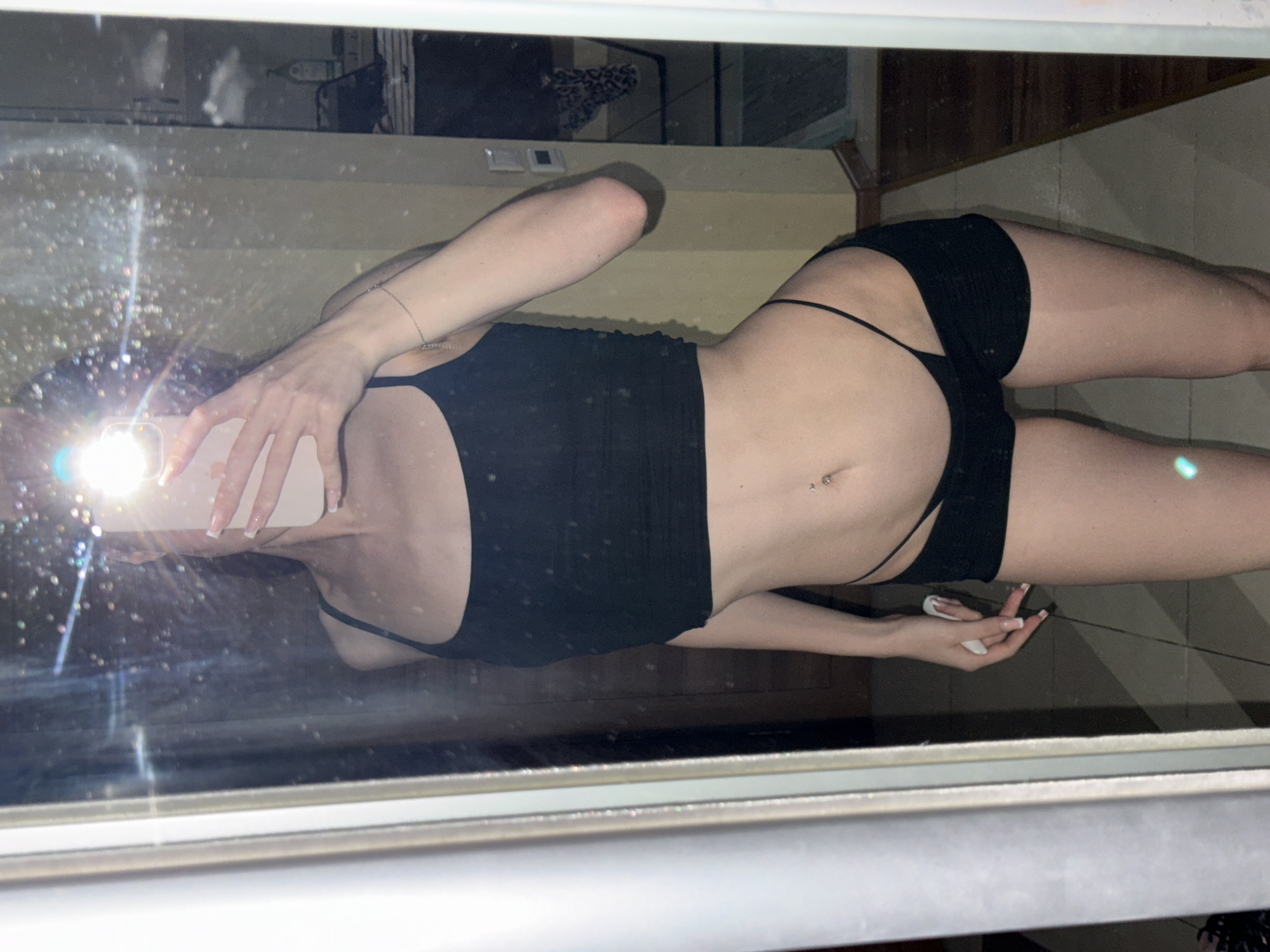 This media file is a mirrored image of a person in a dimly lit room. The subject is a person wearing black lingerie, with a black tank top and black bikini bottoms. They are posing in a way that highlights their midriff and legs, with a belly button piercing visible. The person's hand is holding a smartphone, which is the source of the bright light reflecting in the mirror. The reflection also shows a cluttered background with what appears to be furniture and possibly clothing. The overall impression is that of a casual selfie taken in a personal space, likely a bedroom or bathroom. The lighting is low, with a strong flash from the phone creating a halo effect. There are no discernible signs of the time of day, weather, or specific emotions being conveyed. The location is given as Khimki, Russia, but there are no specific visual cues in the image to confirm this.