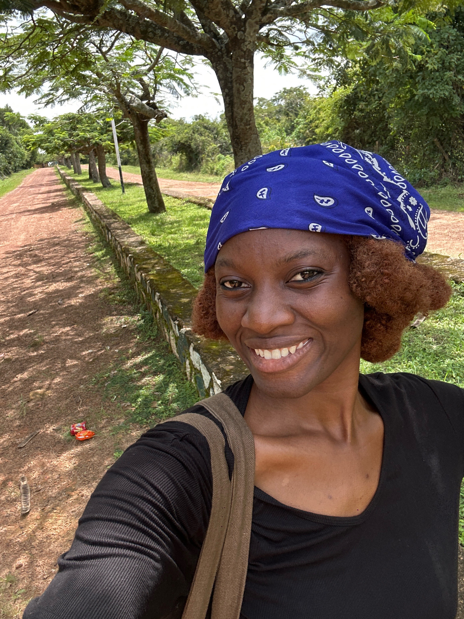 A young Black woman with brown curly hair and a blue bandana smiles at the camera. She is wearing a black top and a brown strap is visible across her shoulder. She is standing outdoors on a dirt path lined with grass and a stone border. Trees are visible in the background, casting shadows on the path. The location is not specified as Jos, Nigeria.