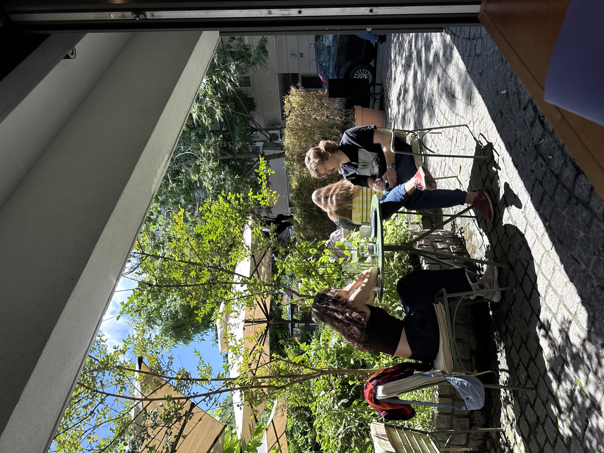 Captured from within an interior space looking out through an open doorway, the image depicts two individuals seated at a small, round, green metal table in an outdoor setting.
One person, facing left, is wearing a black top and dark pants and has long, curly, dark hair. A red and white patterned shirt and a blue bag are draped on the back of the chair.
The other person, facing right, is wearing a black t-shirt with a graphic and blue jeans, paired with red shoes.
Both individuals are sitting on green metal chairs, typical of outdoor cafes.
The table between them has glasses and other items.
The outdoor area appears to be a patio or garden, with trees providing shade and partially obscuring the view of surrounding buildings and parked vehicles. The ground is paved with cobblestones. The shadows cast across the scene indicate it is daytime, with fairly strong sunlight.
Based on visual clues, this outdoor area is likely the Cafe Florija, situated in Belgrade, Serbia.