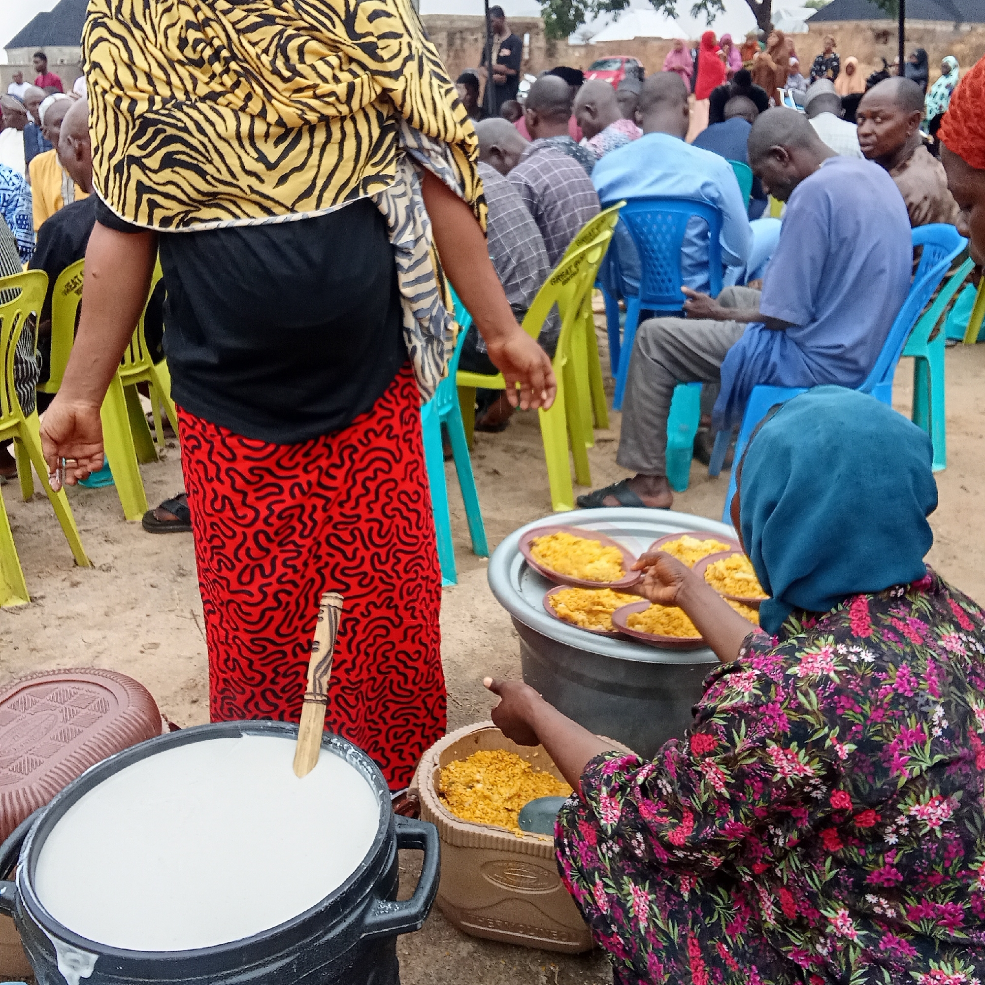 In Doma, Nigeria, an outdoor gathering is underway. Numerous individuals are seated on plastic chairs of green and blue hues. A person stands at the forefront, draped in a vibrant yellow and black zebra-striped shawl over a black top and a red skirt adorned with black squiggles. To their right, a woman in a dark floral print dress and a blue headscarf serves food from trays atop a large container. Beside her sits a black pot filled with a white, porridge-like substance and a wooden spoon sticking out. There are other containers of food, and the setting suggests a communal meal.