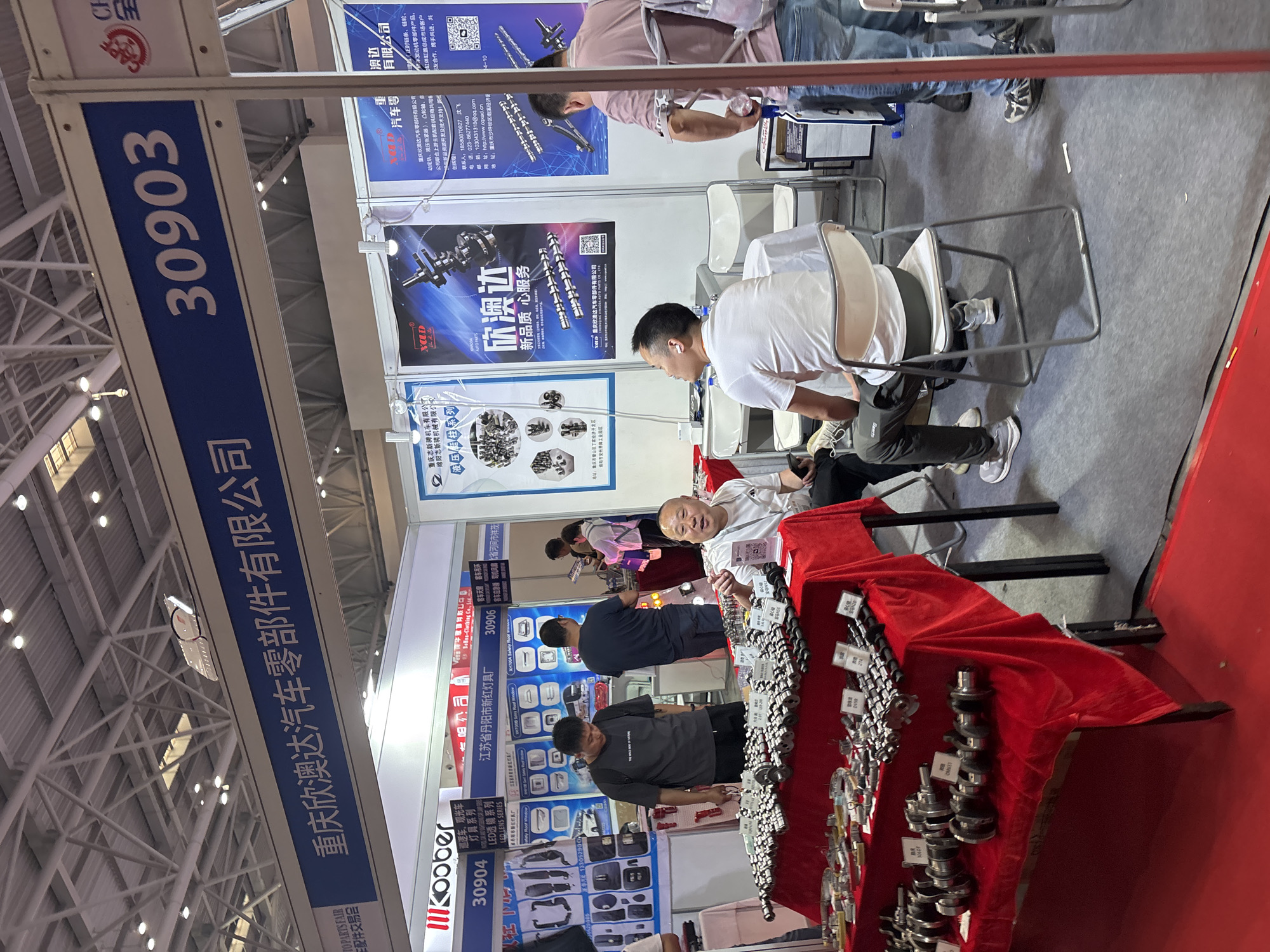 The image shows a trade show booth, possibly in Liangjiang, China, with a sign indicating it belongs to Chongqing Xinaoda Auto Parts Co., Ltd. Booth number is 30903. People are present, including potential customers and booth representatives. The booth displays various car parts, such as camshafts and crankshafts, on a red-draped table. Banners and posters showcase company information and products. Nearby booths belong to Jiangsu Danyang New Red Lantern Factory (30904) and Bollss-Clothing Co., Ltd (30906). A man is seated, engaging with a booth representative.
