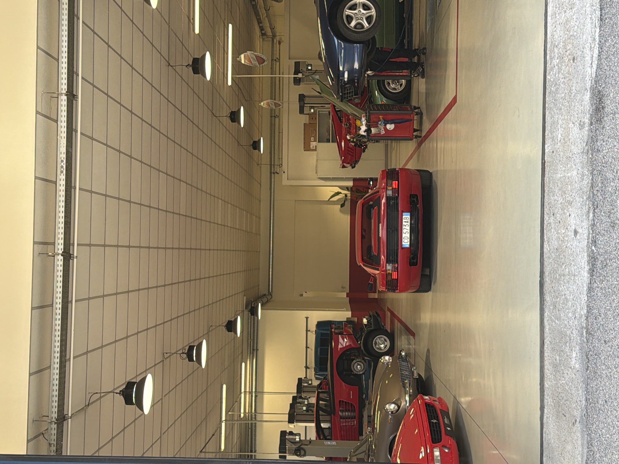 The image depicts the interior of a high-end car workshop in Milan, Italy, specializing in luxury sports cars. The scene is brightly lit by industrial fluorescent lights and overhead pendant lamps. The floor is a polished concrete surface, marked with red lines. In the foreground, a red Ferrari Testarossa is prominently displayed, facing the viewer. To its left, another red Ferrari is partially visible. Further back and to the left, a silver classic car is parked. In the center of the image, a red Ferrari is elevated on a hydraulic lift, its engine exposed, suggesting it is undergoing maintenance. To the right, a blue sports car is also on a lift, with a person in black pants and a dark shirt working on it. Various tool carts and equipment are scattered around the workshop, indicating an active working environment. The overall impression is one of meticulous care and professional expertise applied to prestigious vehicles.