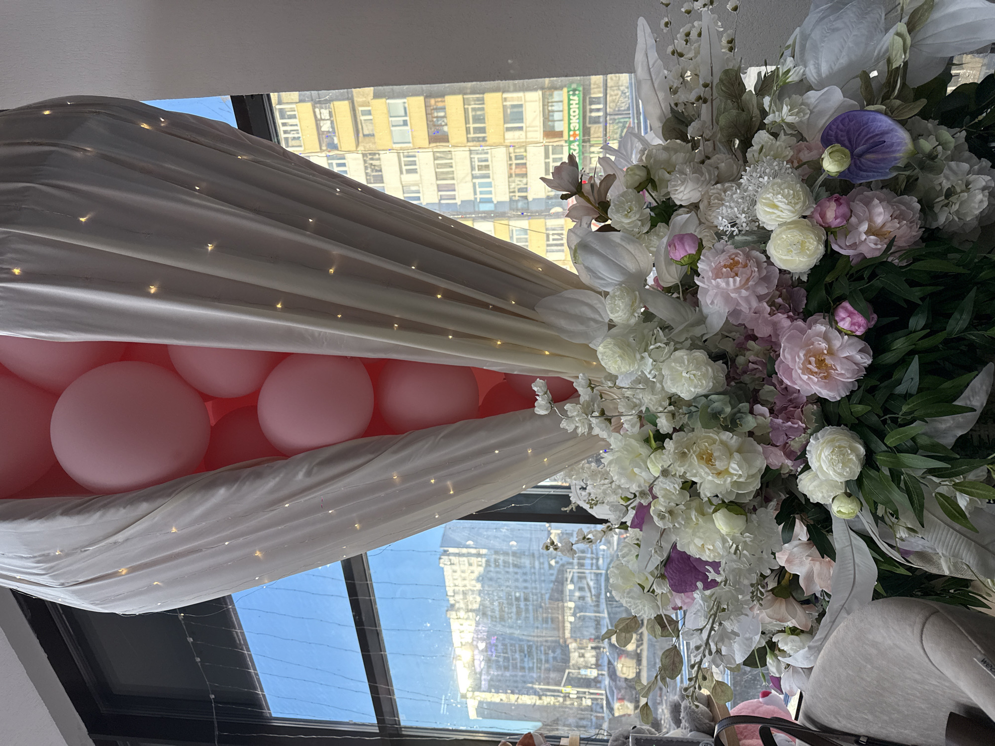 A decorative setting is inside what appears to be a room with large windows offering a view of urban buildings, presumably in Nizhnevartovsk, Russia, judging from the visual context.
The setting features a decorative arrangement of pink balloons nestled beneath a draped white fabric, which is adorned with small, fairy-like lights. The fabric and balloons are positioned near a window.
On the right side of the frame is a lush floral arrangement with white, pink, and purple flowers with green foliage. A part of a gray chair or seat is visible in the lower right corner.
The exterior environment includes buildings with varying heights and facades, visible through the window. The time of day is likely during daylight hours, given the clear sky and light.