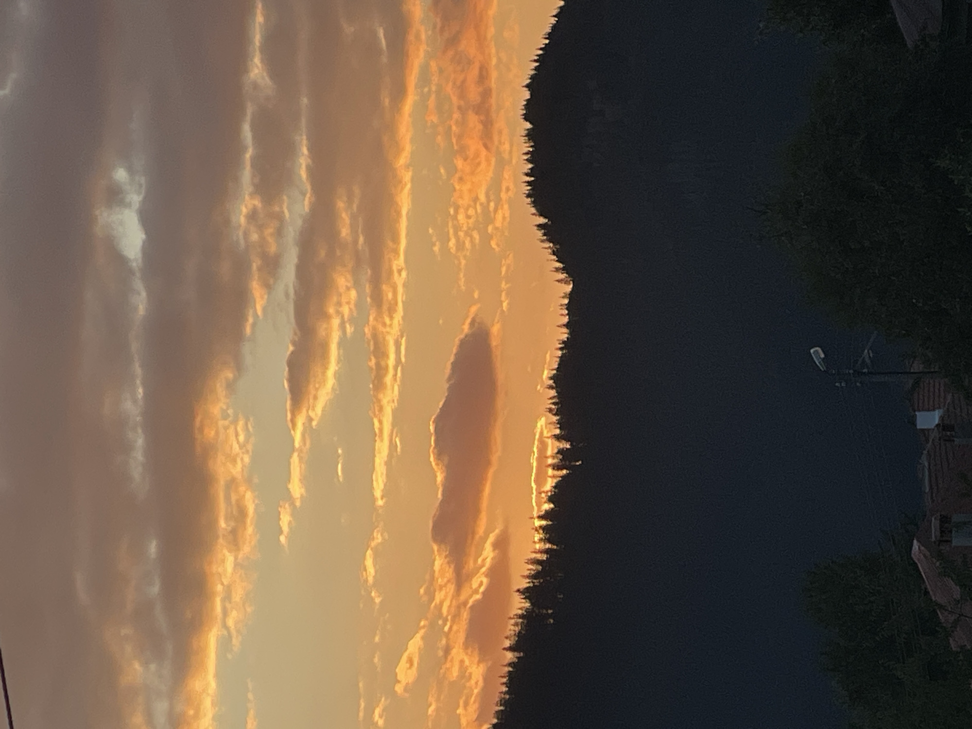 The image depicts a sunset over a mountainous landscape, likely in or near Manastir, Bulgaria. The sky is filled with dramatic clouds colored in vibrant oranges, yellows, and browns, indicating the sun is low on the horizon. A silhouetted mountain range, covered with dense coniferous trees, dominates the middle ground. The dark silhouette contrasts sharply with the brightly lit sky. In the foreground, partially visible rooftops of houses and dense green trees suggest a rural or semi-urban settlement at the base of the mountains. A solitary street lamp is also visible in the lower right quadrant, adding a touch of human infrastructure to the natural scene. No people are visible in the frame.