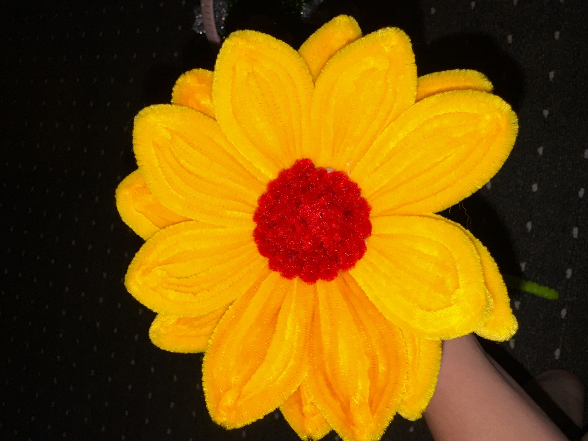 A close-up, top-down shot captures a vibrant, handmade flower crafted from yellow and red pipe cleaners against a dark, textured background. The flower, resembling a daisy or sunflower, features numerous yellow petals radiating from a dense, red center. The texture of the pipe cleaners is visible, giving the petals a fuzzy appearance. A human arm and hand, out of focus, enter the frame from the lower left, holding the stem of the flower, which is a green pipe cleaner. The background appears to be a dark fabric, possibly a carpet or tablecloth, with small, scattered white dots. The lighting is direct, casting subtle shadows and highlighting the plush texture of the pipe cleaner material. No elements in the image suggest a specific location within Chardara, Uzbekistan; the scene appears to be indoors or in a controlled environment.