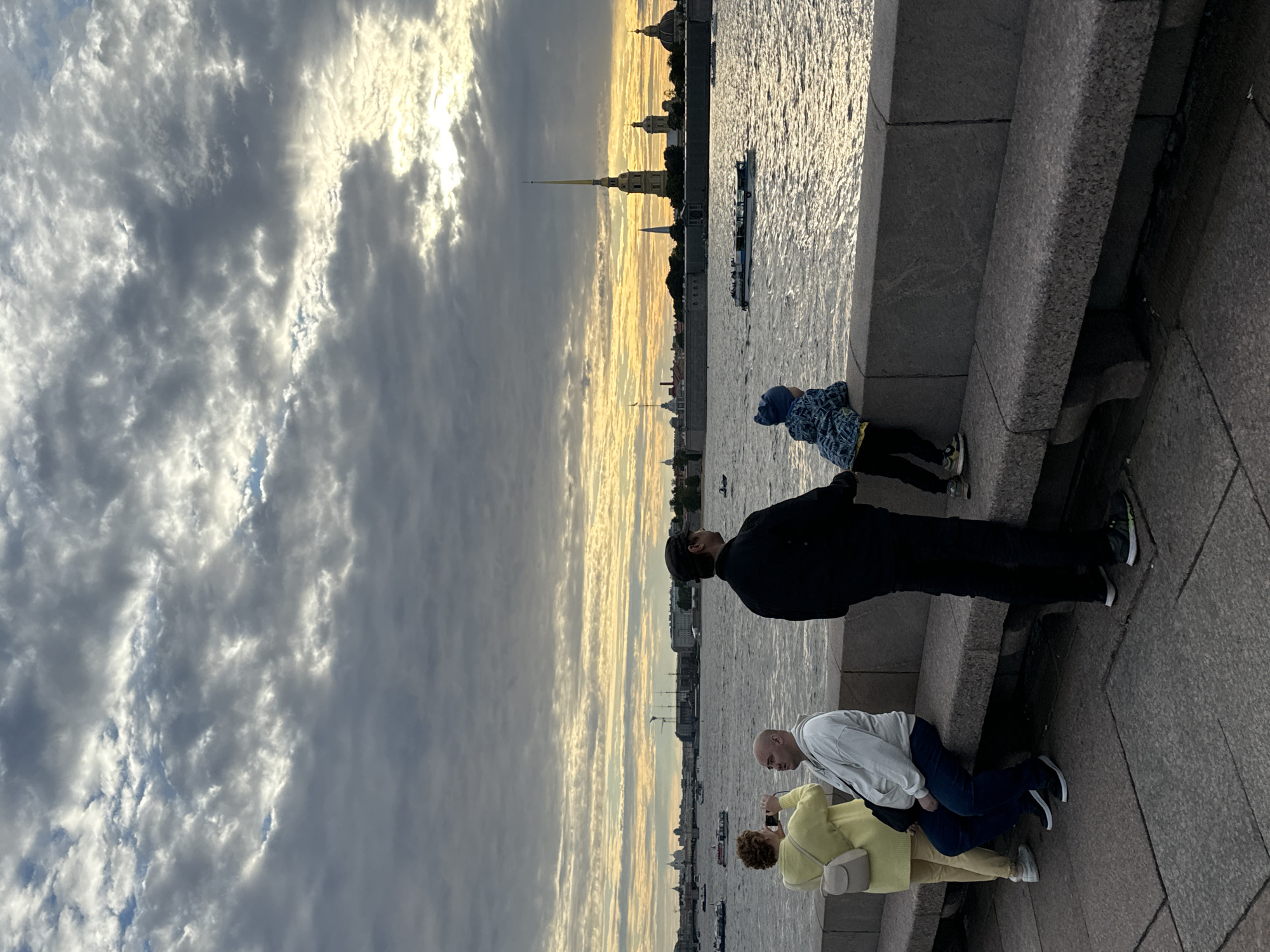 This is a photograph of the Neva River in Saint Petersburg, Russia, at sunset. The sky is filled with dramatic, striated clouds in shades of grey, white, and yellow, indicating the sun is low on the horizon. The river is choppy, reflecting the muted light.
In the foreground, a granite embankment runs along the river. On the embankment, a group of people are visible. On the left, a woman in a yellow sweater and yellow pants is taking a photo with her phone, standing next to a man in a white hoodie and blue jeans. To their right, a man in a black jacket and black pants is looking out at the river. Further to the right, a child in a blue patterned jacket and black pants is leaning over the embankment, looking at the water. A stone bench is placed along the embankment.
Across the river, the cityscape of Saint Petersburg is visible. Dominating the skyline is the spire of the Peter and Paul Fortress. Other buildings with domes and spires are also present, silhouetted against the colorful sky. Several boats are on the river, one in the middle distance appears to be a tour boat. The overall atmosphere is serene and contemplative, with the natural beauty of the sunset contrasting with the historic architecture.
