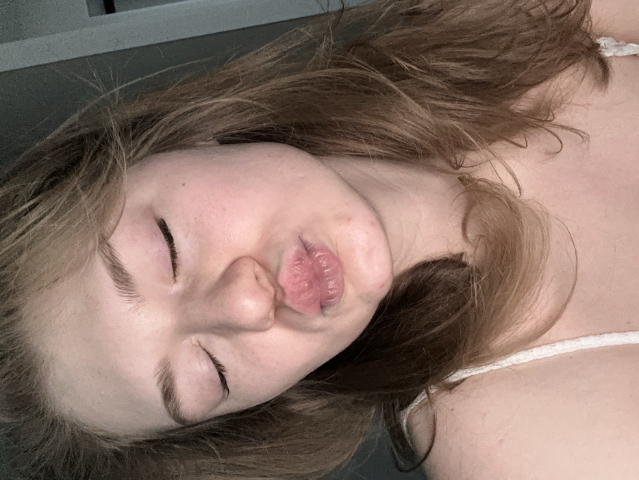 A woman is shown from the chest up, lying on her back with eyes closed. Her fair skin is visible, and she has medium-length brown hair that is slightly wavy. She is making a kissing face. The woman is wearing a white top with thin straps. The background is a blurred combination of grey and indistinct shapes. The overall lighting is soft and diffused.