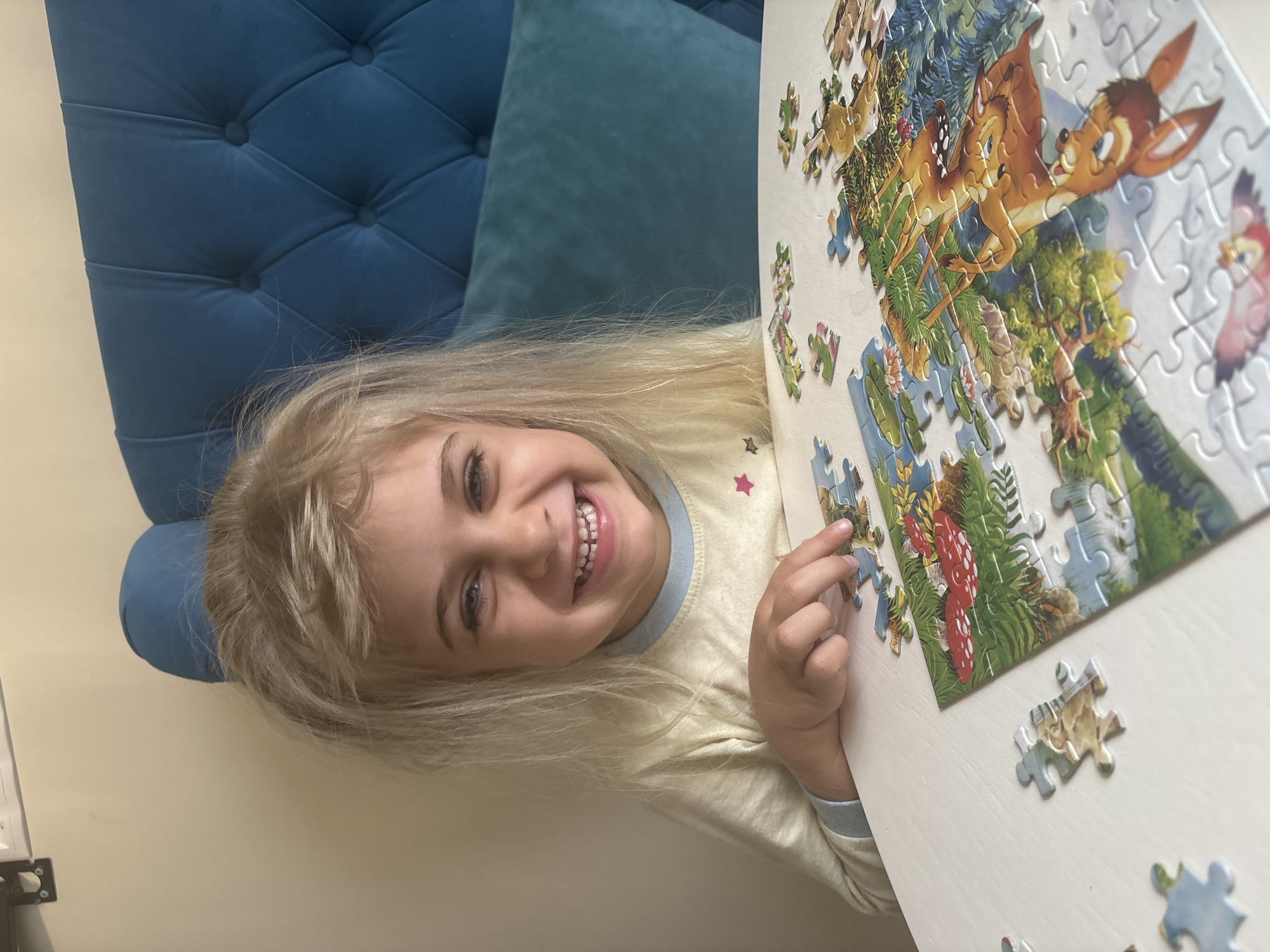 A close-up, slightly tilted shot shows a young girl with light blonde hair and blue eyes smiling at the camera while working on a puzzle. She is wearing a light yellow shirt with a light blue collar and cuffs, adorned with a small pink star. The puzzle depicts scenes from the movie Bambi. It is set on a white table, with a blue tufted chair and pillow visible in the background. The location appears to be indoors, possibly in a home in Unknown, Unknown. The girl is holding a puzzle piece in her hand, suggesting active engagement with the puzzle.