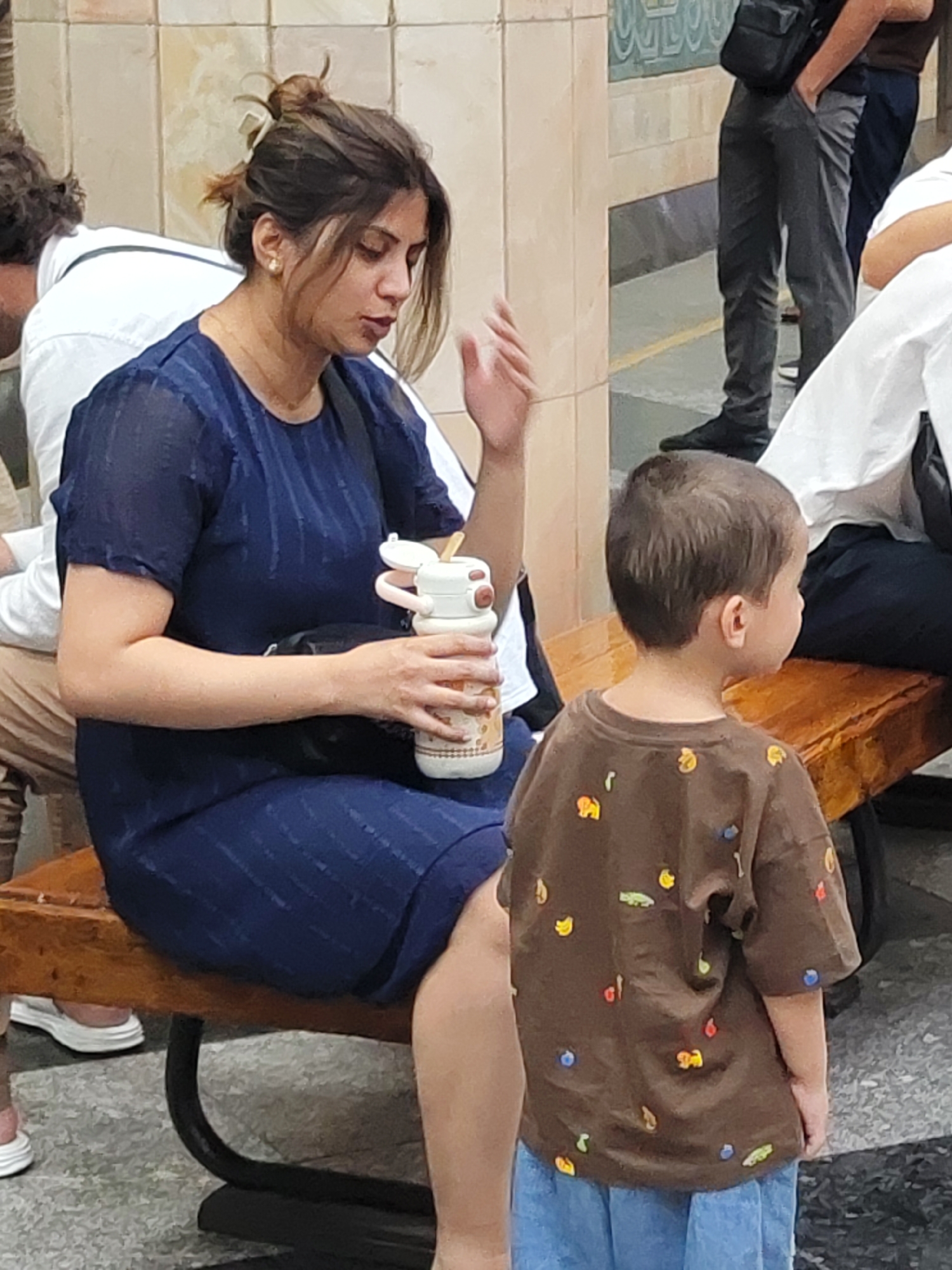 A woman in a dark blue dress sits on a wooden bench in a subway station, holding a patterned water bottle. Next to her, a young boy in a brown shirt adorned with colorful shapes stands and looks away. The bench is situated on a patterned floor, with a tiled wall in the background. The scene suggests a public transportation setting, likely in Tashkent, Uzbekistan, given the context of the original request.